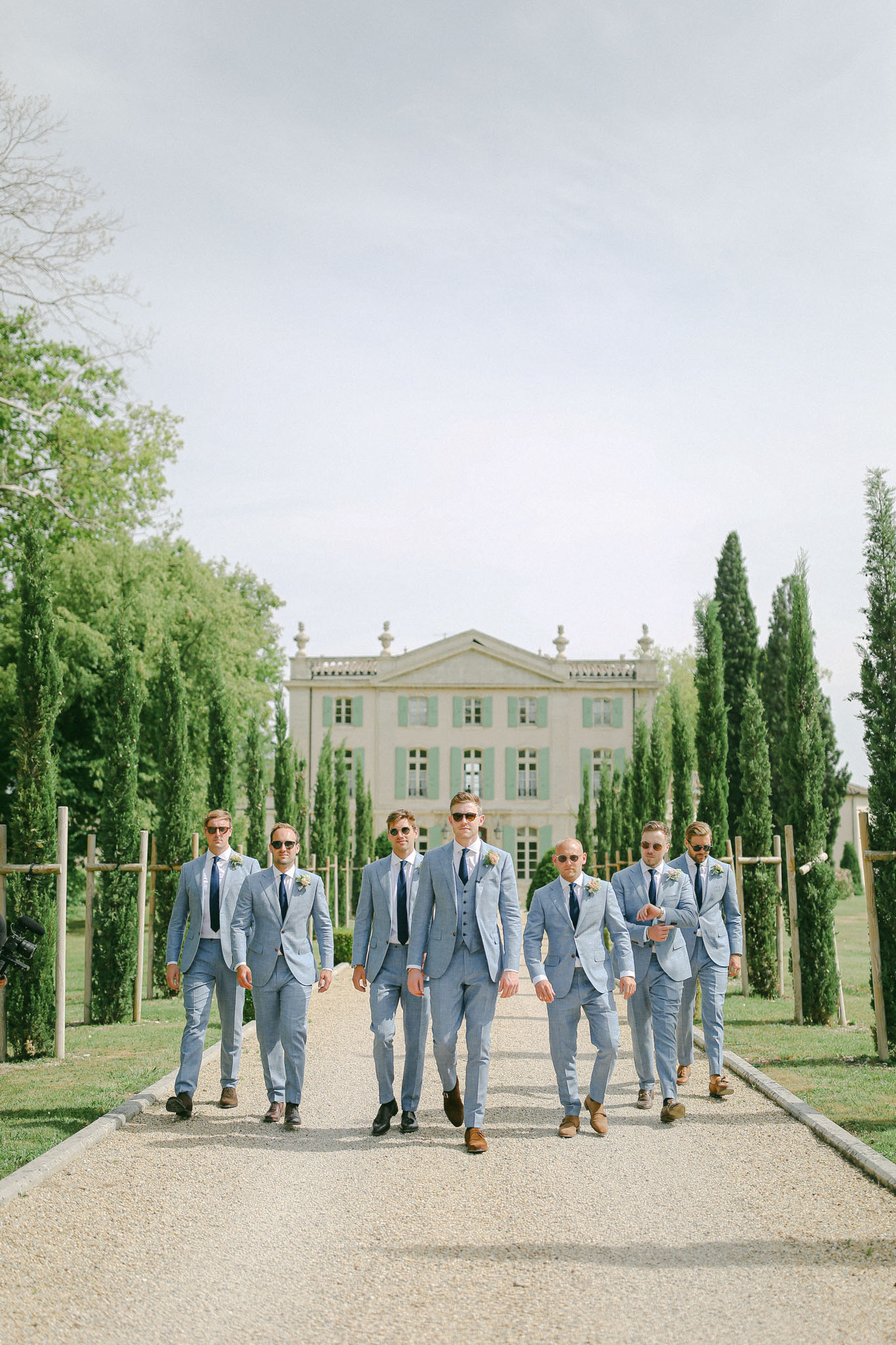 Groom and six groomsmen in light blue suits walking down a gravel allee toward the camera at a French chateau