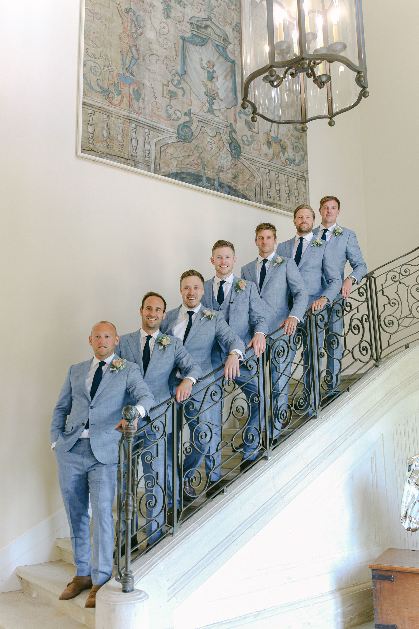 Six groomsmen in light blue suits on chateau interior staircase with wrought-iron banister classical mural and chandelier