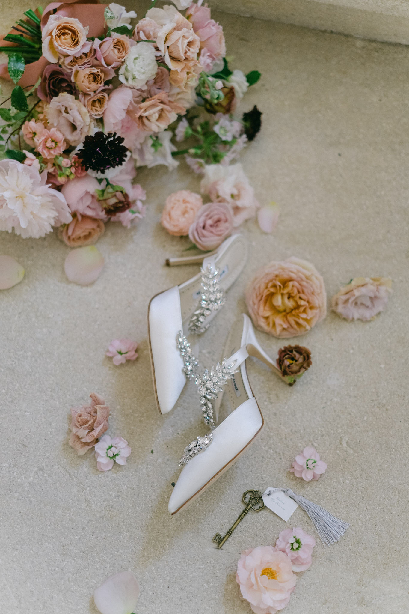 White satin crystal-embellished bridal heels arranged with blush pink and peach roses and antique key on stone surface