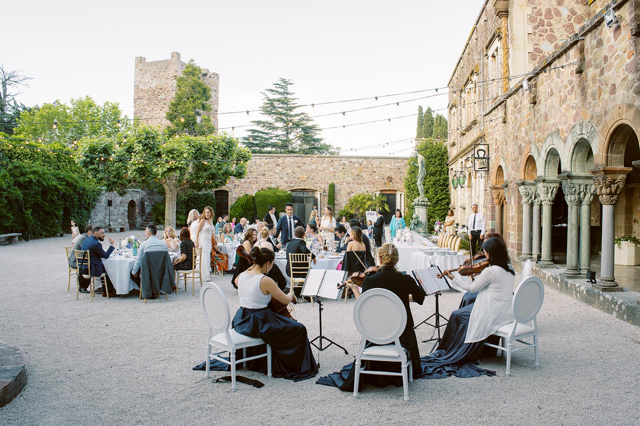 An outdoor wedding reception dinner is underway in the courtyard of a medieval stone château, with approximately 30–40 guests seated at round tables draped in white linen and dressed with gold chiavari chairs. In the foreground, a string quartet of three musicians wearing navy skirts and white tops performs from music stands, seated on white round-back chairs. The bride in a white gown and groom in a dark navy suit are visible standing among the guests mid-reception. Table centerpieces include blue and white floral arrangements. Overhead, warm Edison bulb string lights are strung across the courtyard between trees and the château walls. The venue features a Romanesque stone arcade with carved column capitals along the right side, a medieval tower in the background, and a classical statue visible in the mid-ground. Wide shot taken from a slightly elevated angle capturing both the musical entertainment and the dining guests together. Potential venue feature image.