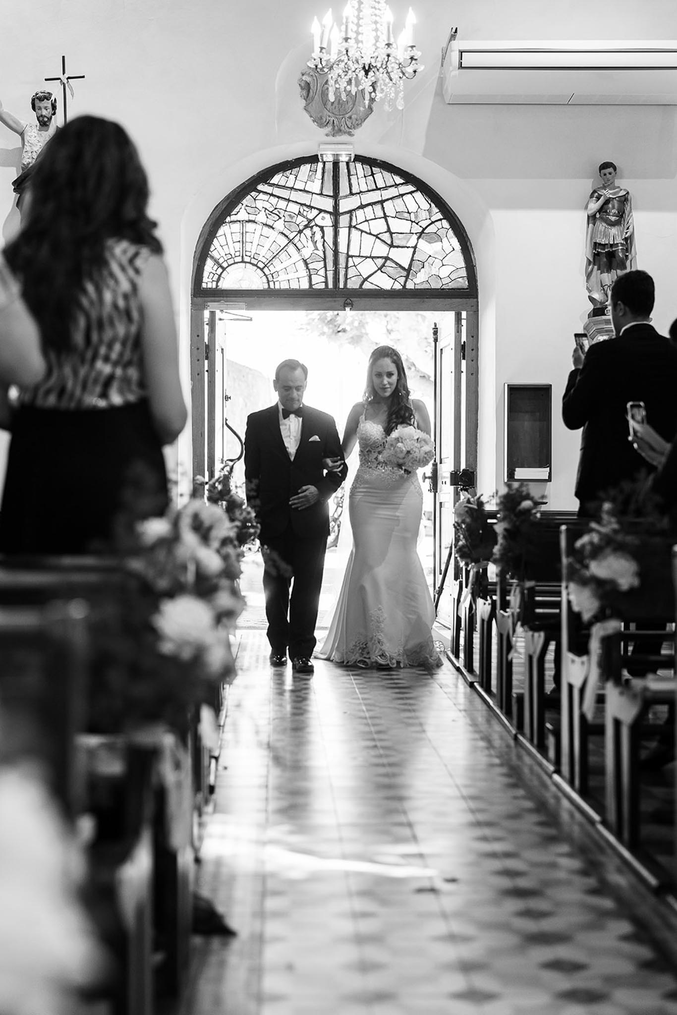 This black-and-white image captures the bride walking down the aisle of a chapel, escorted by a man in a tuxedo, likely her father. The bride wears a fitted mermaid-silhouette gown with lace appliqué along the hem and bodice, and carries a rounded bouquet of light-toned flowers. The chapel interior features wooden pews lined with floral arrangements, a patterned tile floor, religious statues mounted on the walls, and a crystal chandelier overhead. The entrance behind them is framed by a large arched stained-glass window that backlit the pair, creating strong contrast between the bright doorway and the darker interior tones. Guests seated in the pews and standing along the sides are visible, with at least one guest photographing the moment on a smartphone. The composition is a mid-wide shot taken from ground level near the front of the aisle, with a blurred female guest in the foreground adding depth.