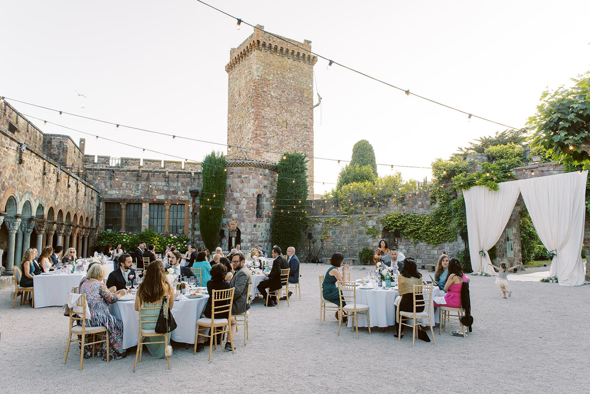 An outdoor wedding reception dinner is underway in the courtyard of a medieval stone castle with a prominent crenellated tower. Approximately 40–50 guests are seated at round tables covered with white linen tablecloths and set with glassware, arranged across a gravel courtyard. Seating uses gold Chiavari chairs throughout. String lights with Edison bulbs are strung across the courtyard, and a white draped fabric arch or curtain installation is positioned to the right side of the space. Table centerpieces include blue and white floral arrangements. Guests are dressed in a mix of cocktail attire including teal, fuchsia, and dark formal wear. A child is visible running near the drape installation on the right. The setting features ivy-covered walls, cypress trees, and a Romanesque arcade along the left wall, contributing to a classic, historic venue aesthetic. Wide shot taken from a slightly elevated angle. Potential venue feature image.