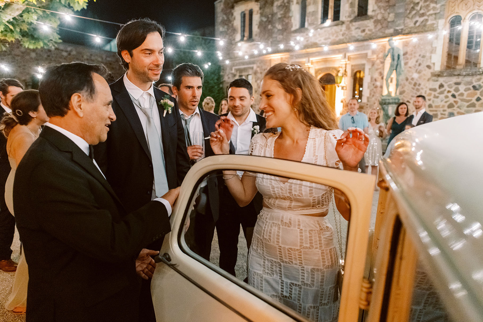 Bride and groom beside vintage cream Fiat 500 during send-off under string lights at chateau courtyard