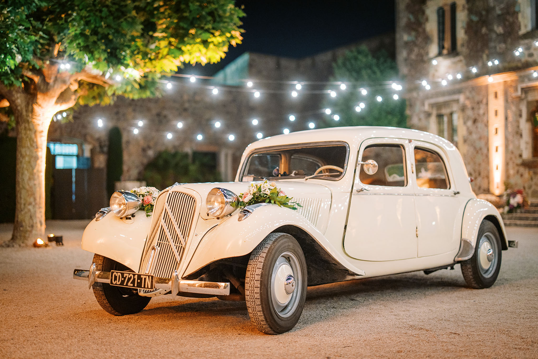 A vintage cream Citroën Traction Avant, decorated with a floral arrangement of blush pink roses and greenery on the hood, is parked in a courtyard at night. The setting is an outdoor courtyard of a French stone building, with globe string lights strung overhead creating warm ambient lighting. No people are visible; the shot focuses entirely on the decorated wedding car as a detail/transport element. The overall styling is classic French vintage, with the warm light from the string lights and uplighting on the stone facade contributing to the evening atmosphere.