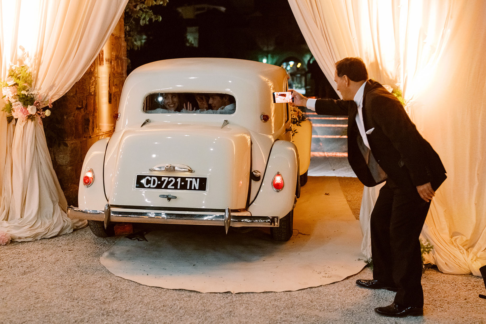 Couple waving from vintage cream car at night venue entrance framed by draped curtains and pink rose arrangements