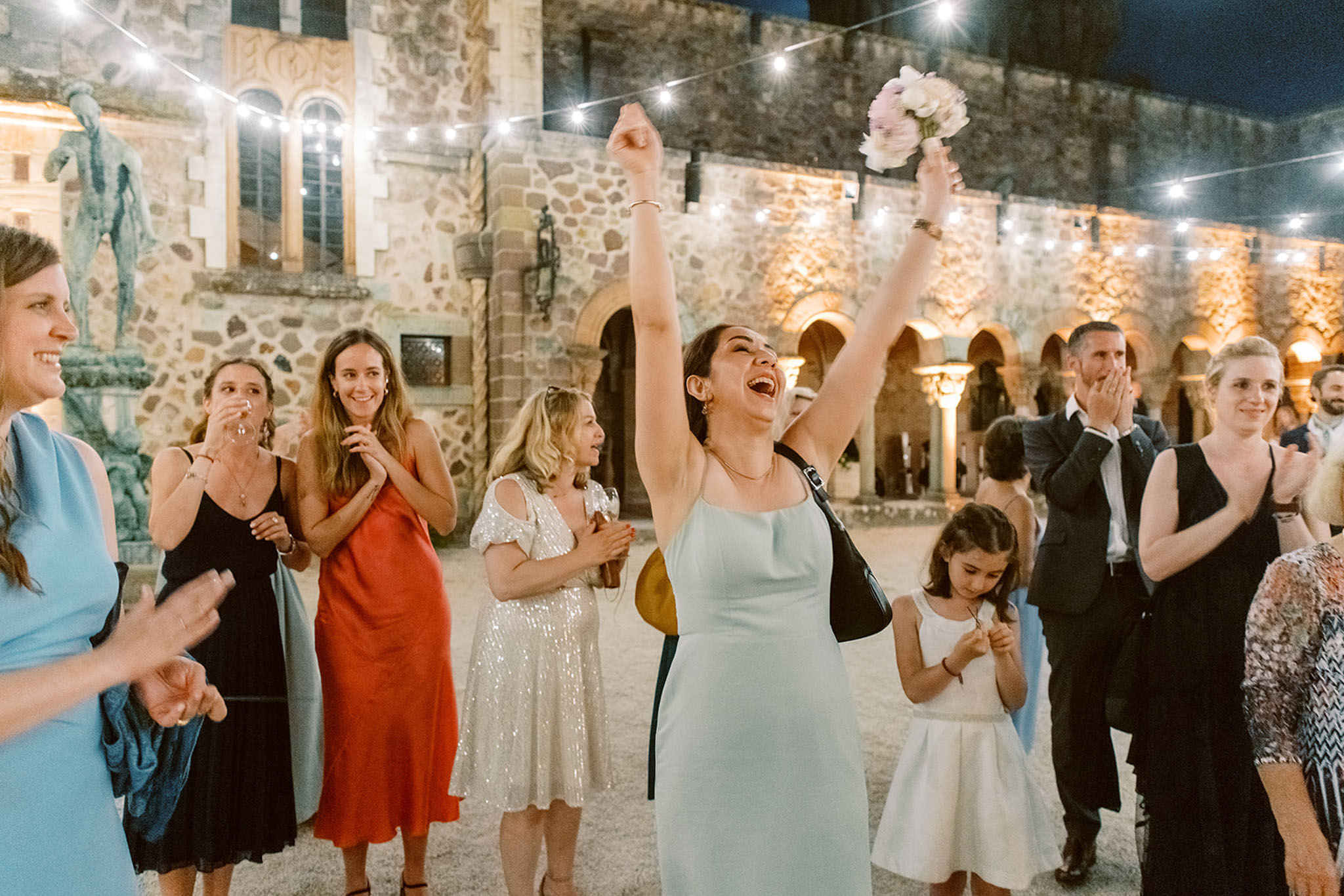 Woman in mint dress catches peony bouquet as guests celebrate under string lights at stone courtyard
