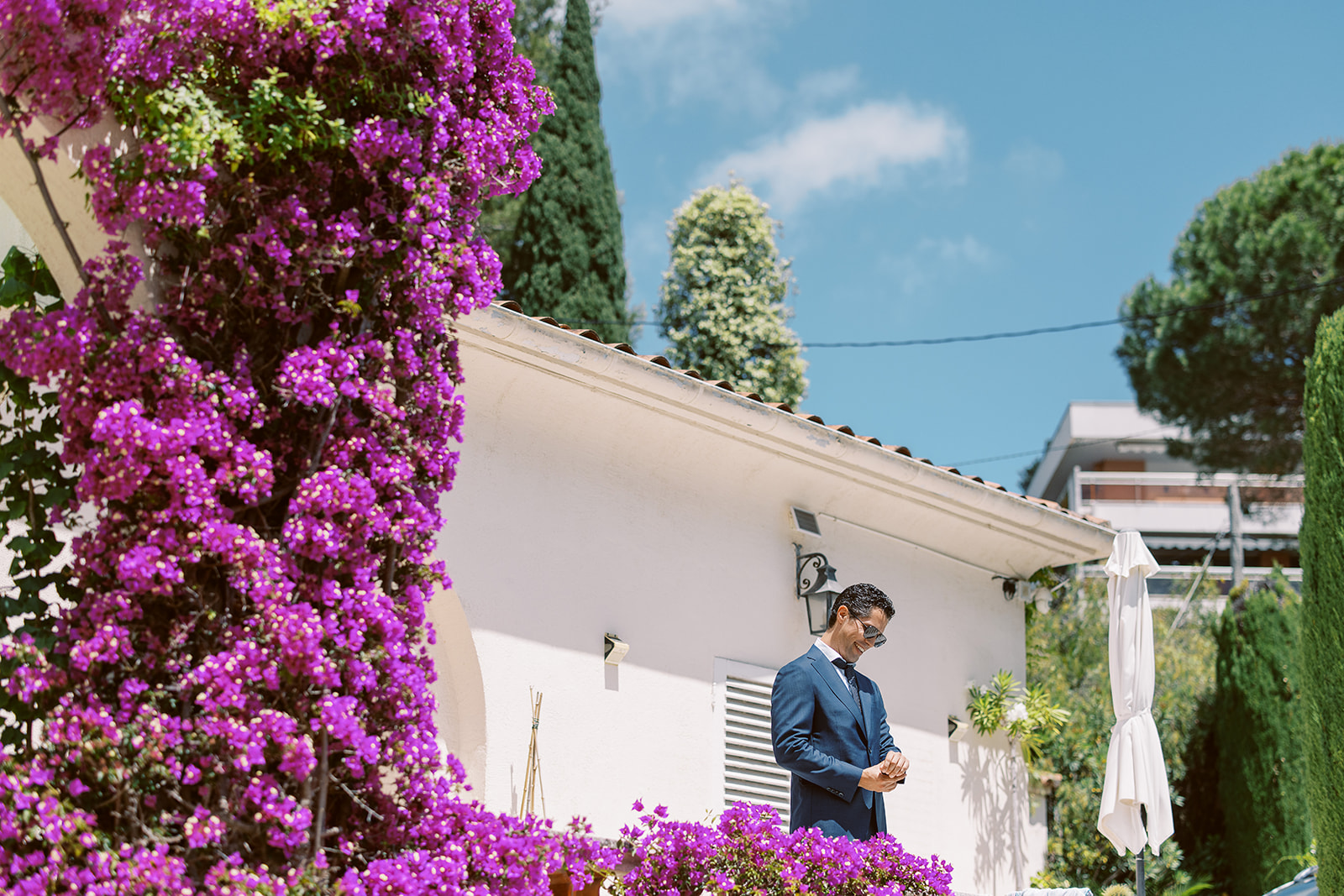 A groom stands outdoors in front of a white-rendered building with a terracotta tile roof, adjusting his cufflinks or jacket while looking down. He is wearing a navy blue suit with a tie and sunglasses pushed down his nose. The foreground is framed by a large bougainvillea vine covered in vivid magenta-purple blooms, which occupies the left side of the frame and draws the eye toward the groom in the mid-ground. A closed white parasol is visible to the right. The composition is a medium environmental portrait taken from a slight distance, using the flowering vine as a natural foreground frame against the white building facade.