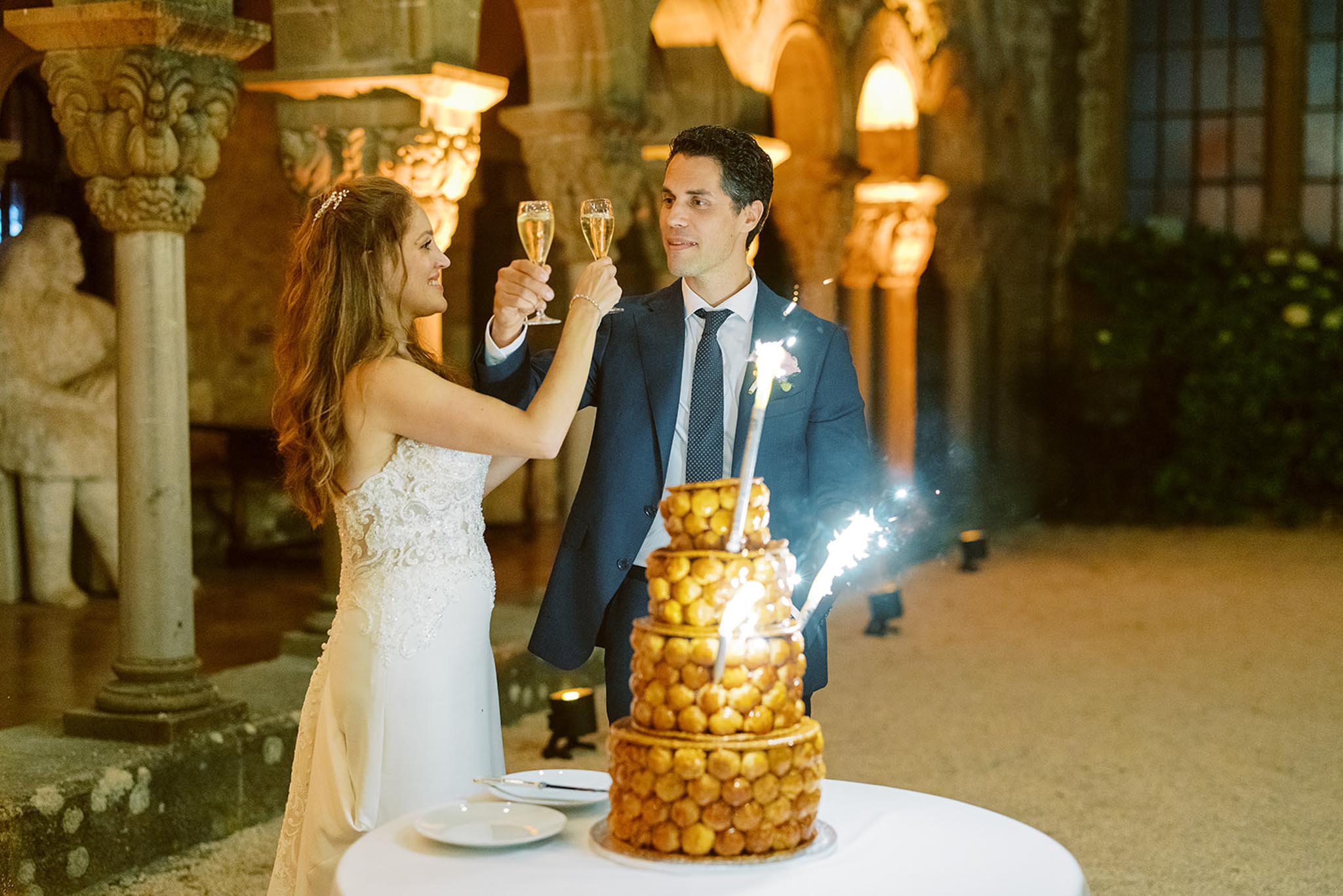 The bride and groom toast with champagne flutes beside a traditional French pièce montée (croquembouche), which is topped with active sparklers. The bride wears a fitted white lace strapless gown with a delicate hair accessory and a pearl bracelet, while the groom wears a navy suit with a navy dotted tie and a small pink floral boutonnière. The setting is an indoor space featuring ornate stone columns, sculpted statues, and warm amber uplighting that creates a golden glow across the scene. The portrait-style shot is taken at medium range, with the croquembouche prominently positioned in the foreground on a white-clothed table.