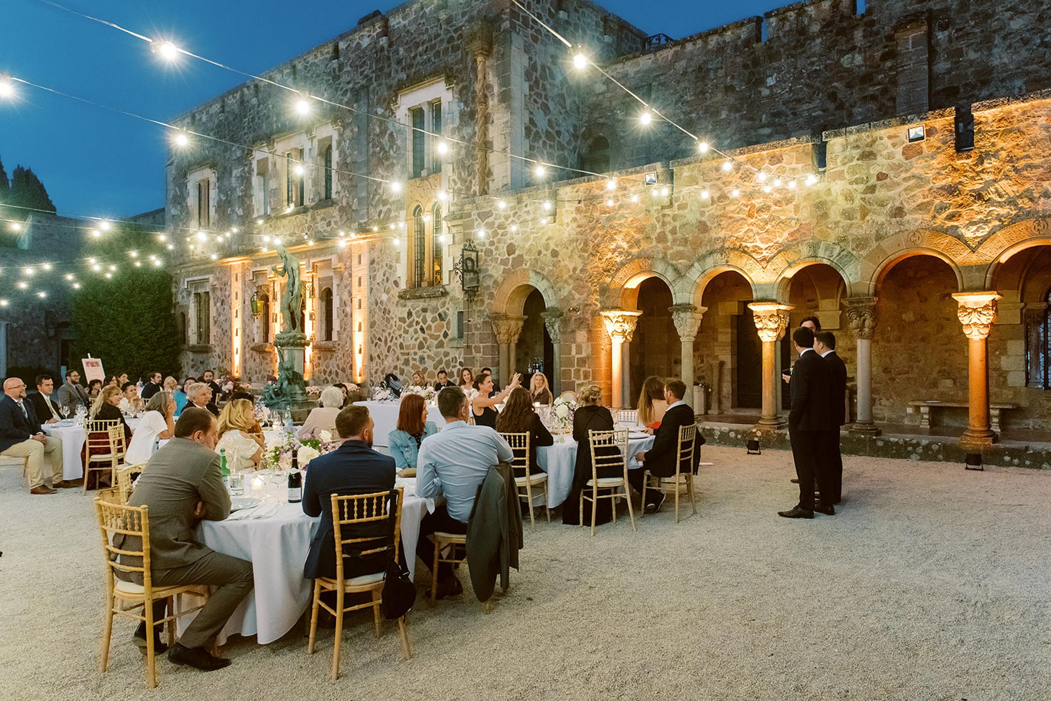 50 guests at round tables under fairy lights in Romanesque arched colonnade courtyard at dusk