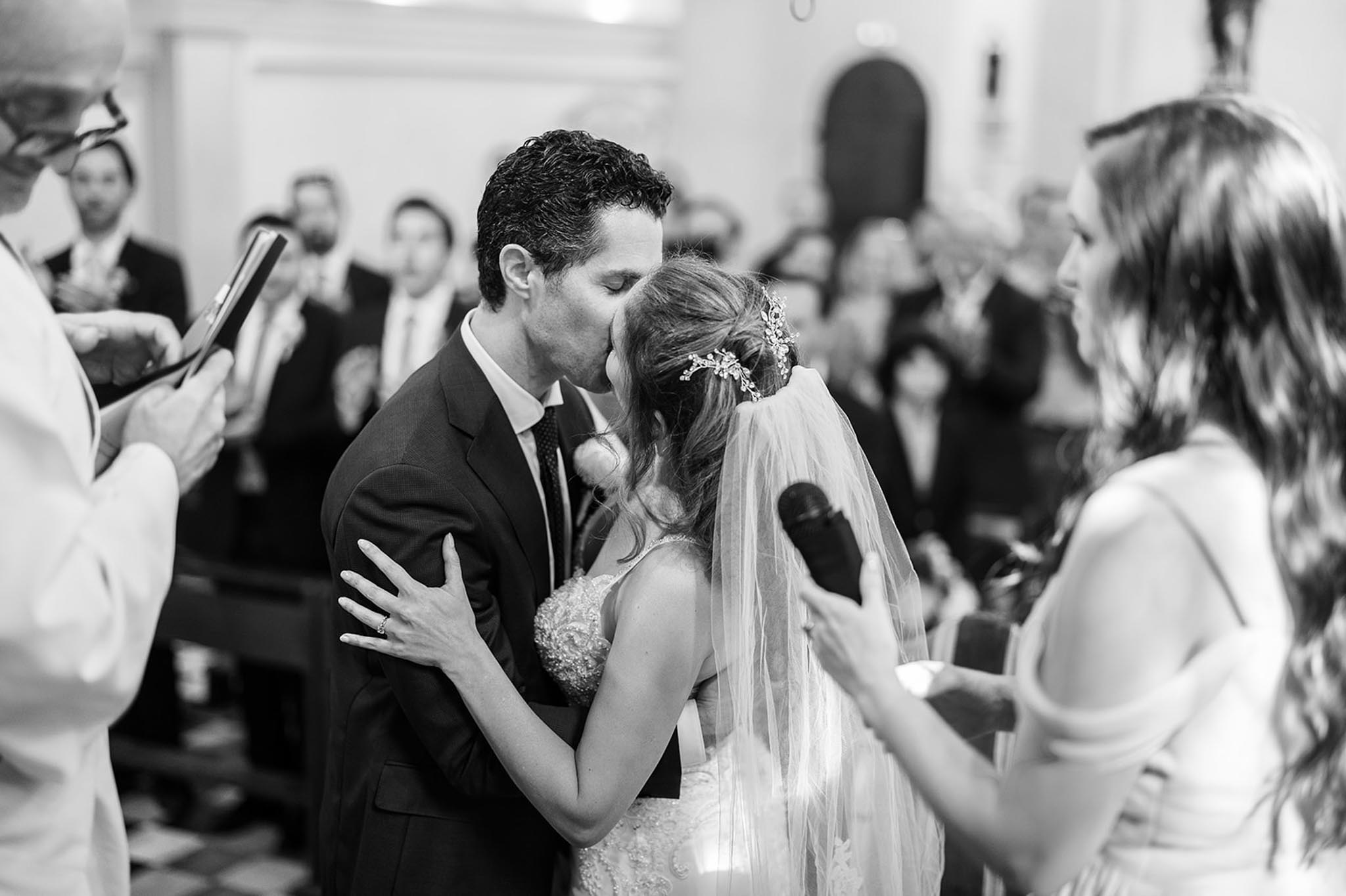 This black-and-white image captures the first kiss moment during an indoor religious ceremony, likely in a chapel or church, with white plastered walls and an arched doorway visible in the background. The groom wears a dark suit with a tie and a boutonnière, while the bride wears a lace-detailed dress, a long tulle veil, and an ornate floral hairpiece. A bridesmaid in a sleeveless dress stands to the right holding a microphone, and an officiant in white robes is partially visible to the left holding a book. A full congregation of seated guests is visible in the background, with several dozen attendees looking on.