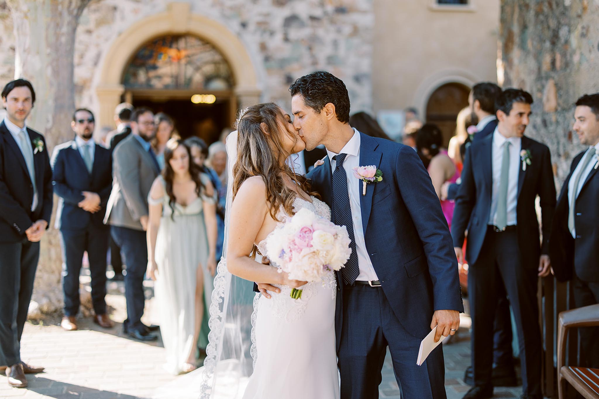 First kiss outside stone chapel with blush peony bouquet and bridesmaid in sage dress
