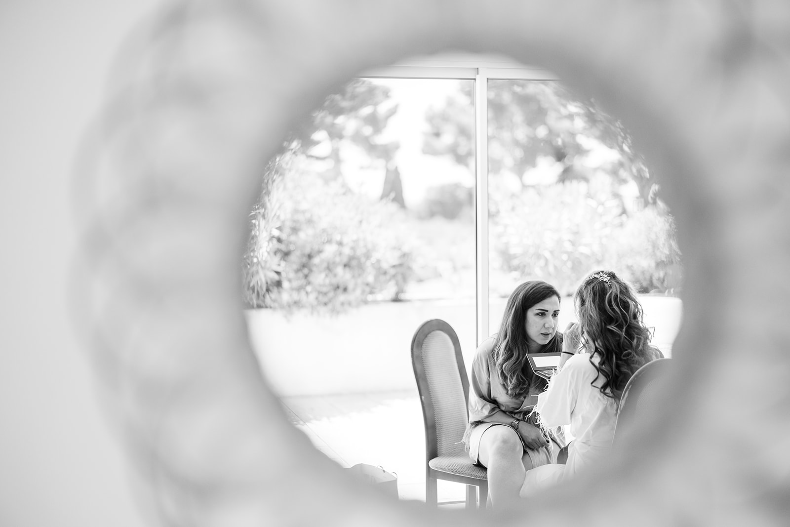 A black-and-white getting-ready scene captured through the circular opening of a mirror, creating a framed vignette effect with soft bokeh on the edges. Two women are seated indoors near a large floor-to-ceiling window with garden greenery visible outside; one woman appears to be a bridesmaid or friend dressed in casual attire holding what looks like a small gift box, while the other, likely the bride, wears a white robe and a floral hair accessory with curled hair. The two are in close conversation, facing each other. The image uses high contrast with bright whites from the window light and soft mid-tones, shot as a creative medium shot using the mirror as a compositional framing device.