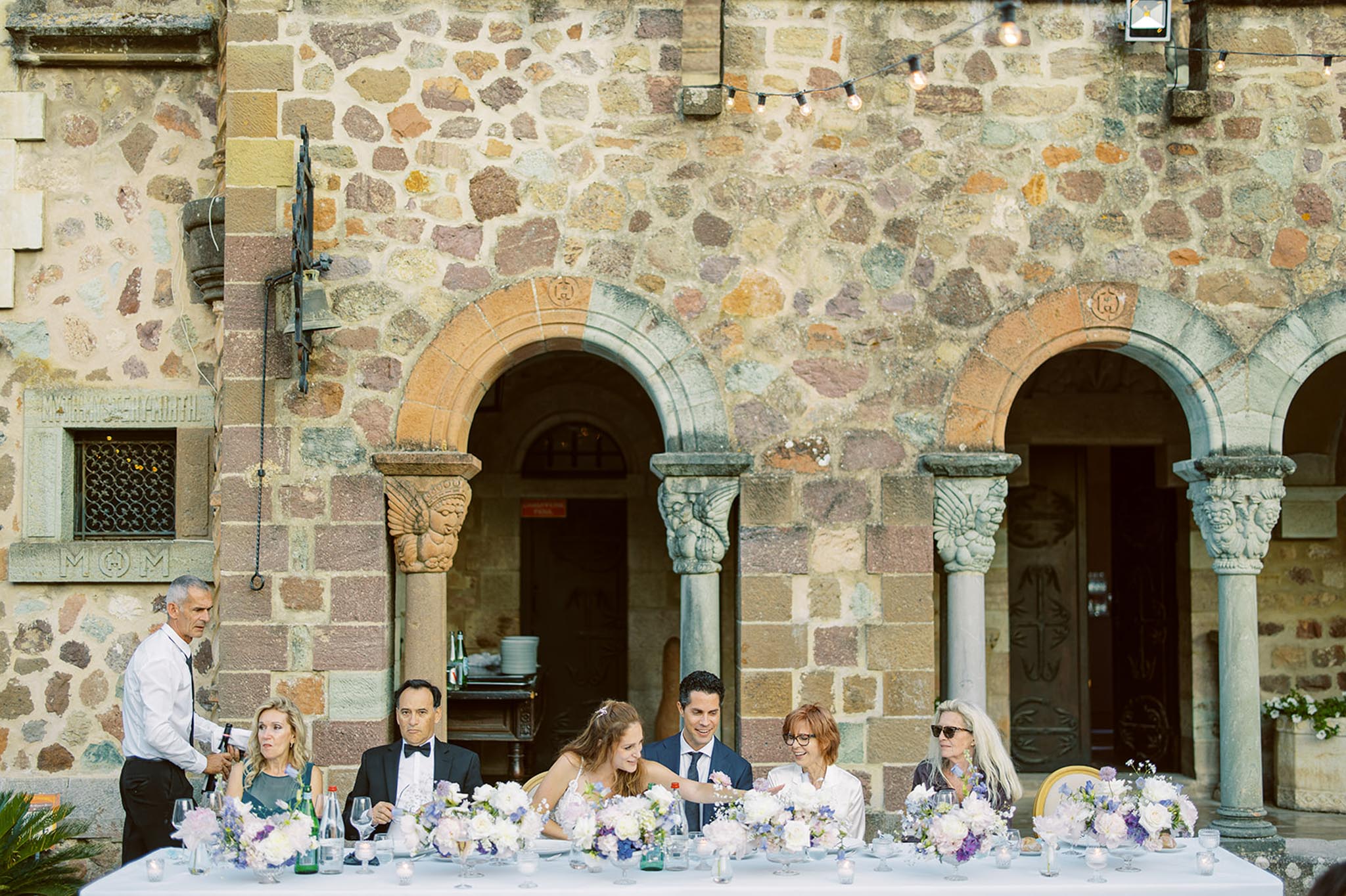 An outdoor wedding reception dinner is taking place in a courtyard against the facade of a historic stone building featuring Romanesque-style arched colonnades with ornately carved capitals and decorative stonework. The head table seats approximately six guests plus the couple — the bride is in a white spaghetti-strap gown and the groom in a navy suit — along with a man in a black tuxedo and several women in evening attire, all engaged in conversation. A member of the catering staff in a white shirt and black tie stands to the left operating an audio device. The white linen-covered table is decorated with multiple low floral arrangements in white, lavender, and soft purple tones featuring what appear to be peonies, hydrangeas, and delicate filler blooms, alongside votive candles and glassware. Edison bulb string lights are strung across the top of the frame, and the wide-shot composition captures both the table setting and the full architectural backdrop. Potential venue feature image.