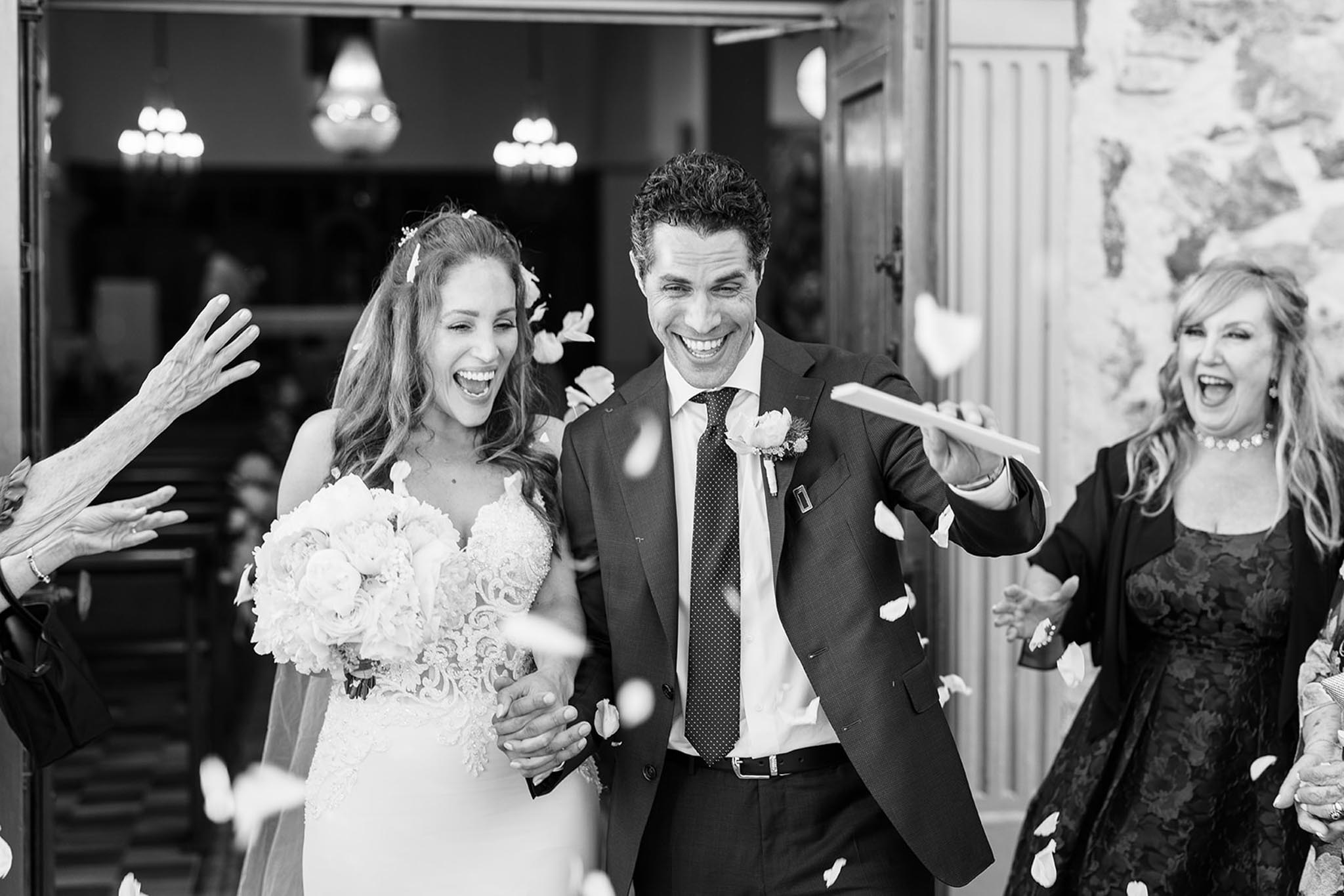 Couple laughs exiting church through wooden doors in shower of flower petals in high-contrast B&W
