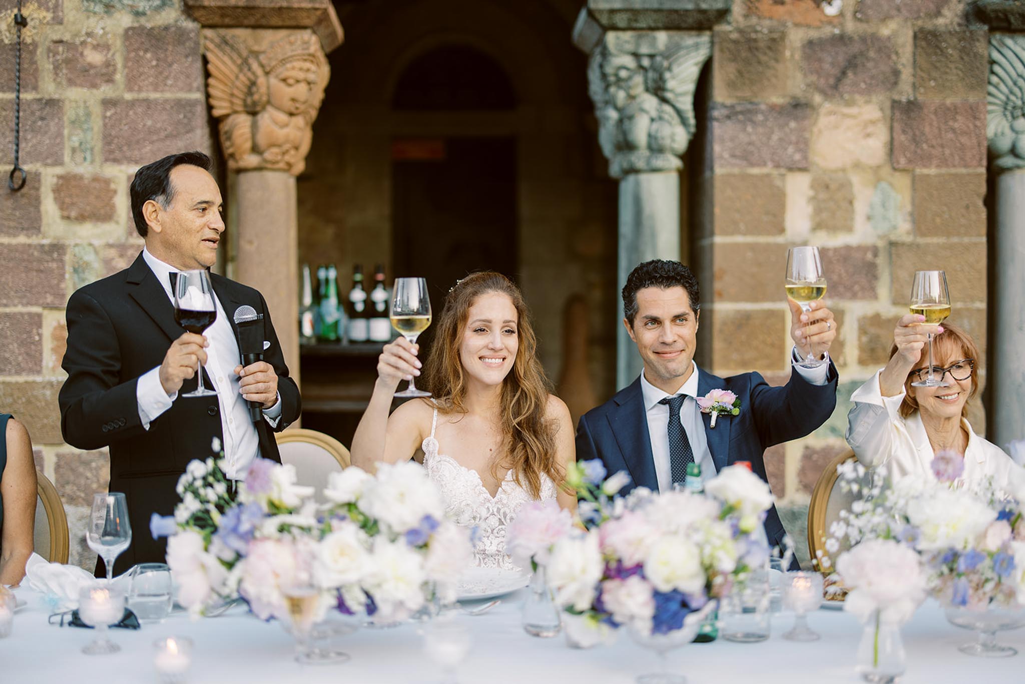 Wedding speech and toast at head table with floral centerpieces before Romanesque stone columns