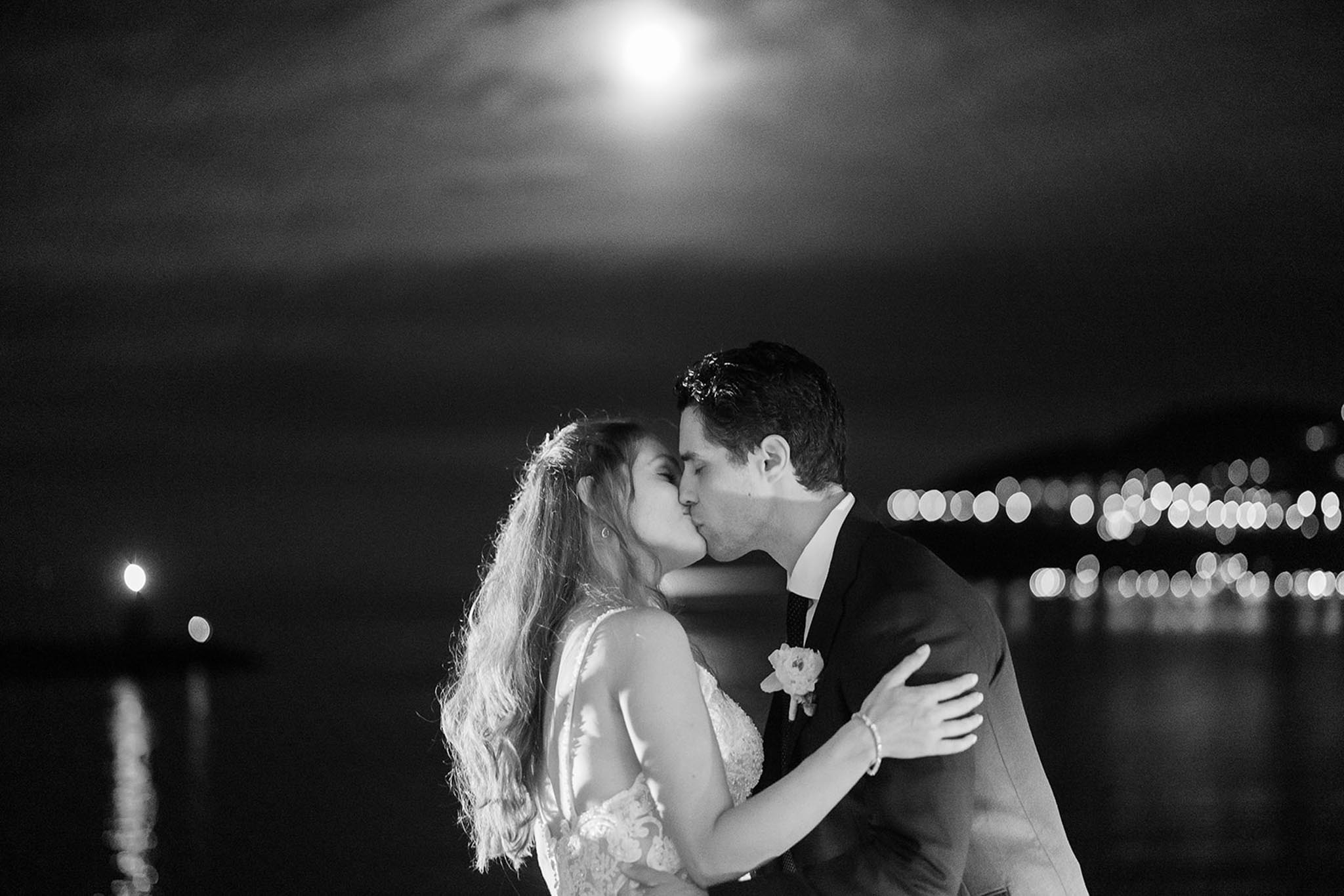 Black-and-white nighttime portrait of bride and groom kissing by waterfront with bokeh lights reflected on water