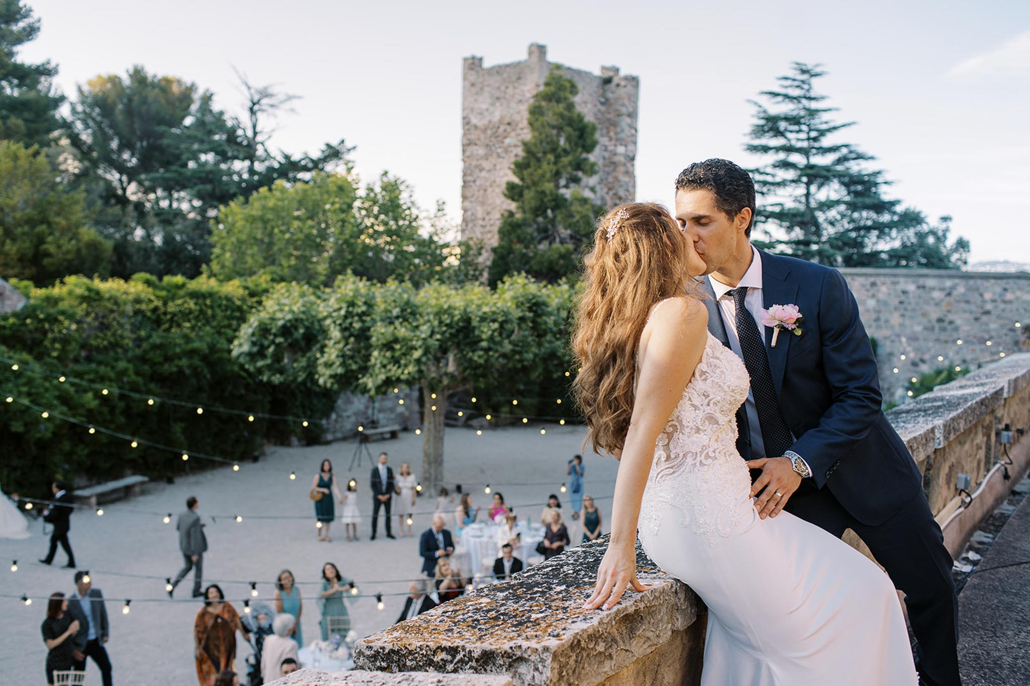 Bride and groom kissing on stone parapet above courtyard cocktail hour with festoon lights and medieval castle tower