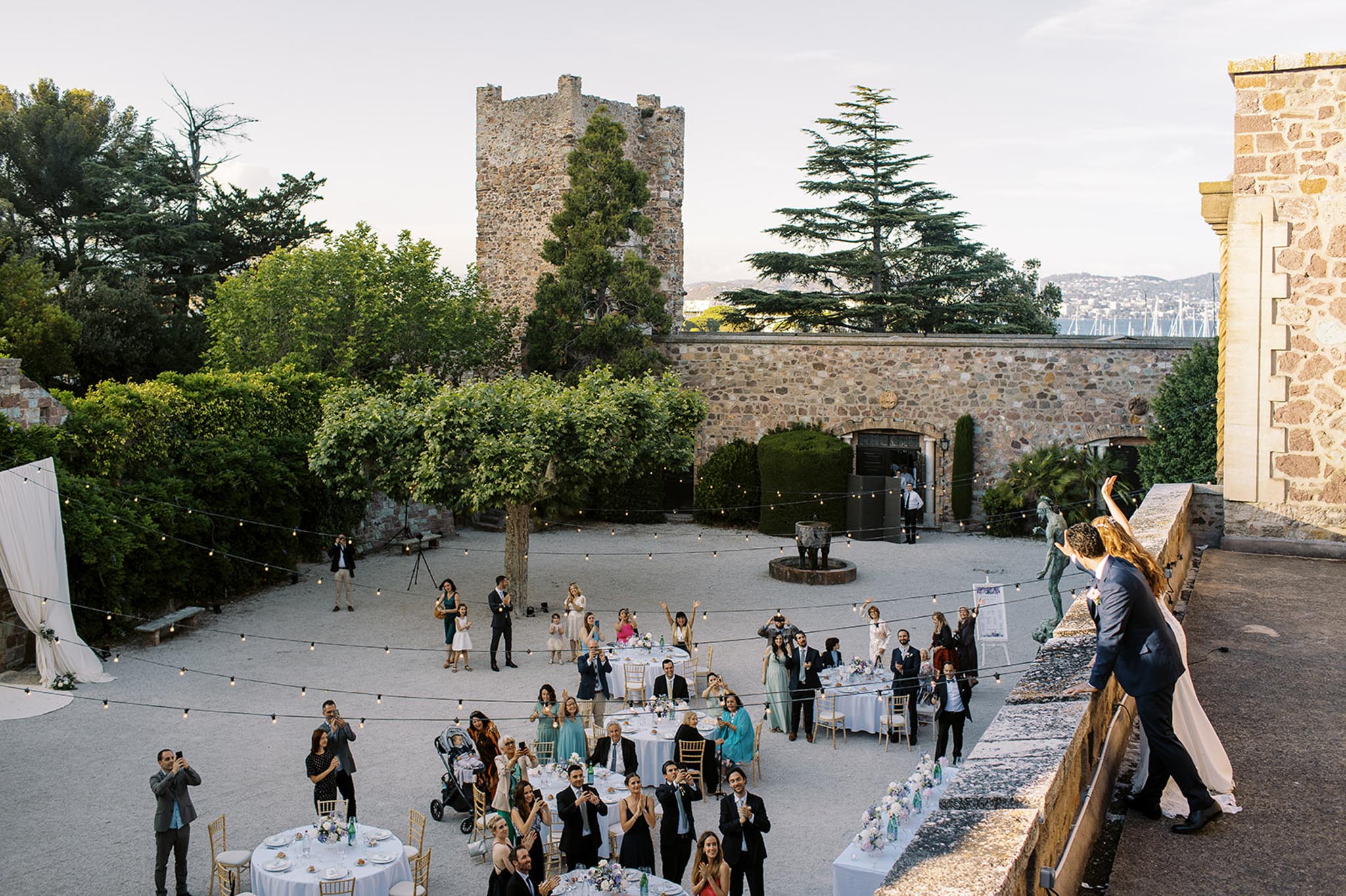 An outdoor wedding reception is taking place in the courtyard of a historic stone château, with a medieval tower and ancient stone walls visible in the background. The bride in a white gown and groom in a navy suit stand on an elevated terrace or balcony to the right, waving to approximately 50 guests gathered below around round tables dressed in white linen with gold Chiavari chairs and small floral centerpieces in soft pastel tones. Festoon bulb string lights are strung across the entire courtyard, creating a warm ambient glow at dusk. The wide-angle elevated shot captures the full layout of the reception space, with guests cheering, raising glasses, and photographing the couple from below. Potential venue feature image.