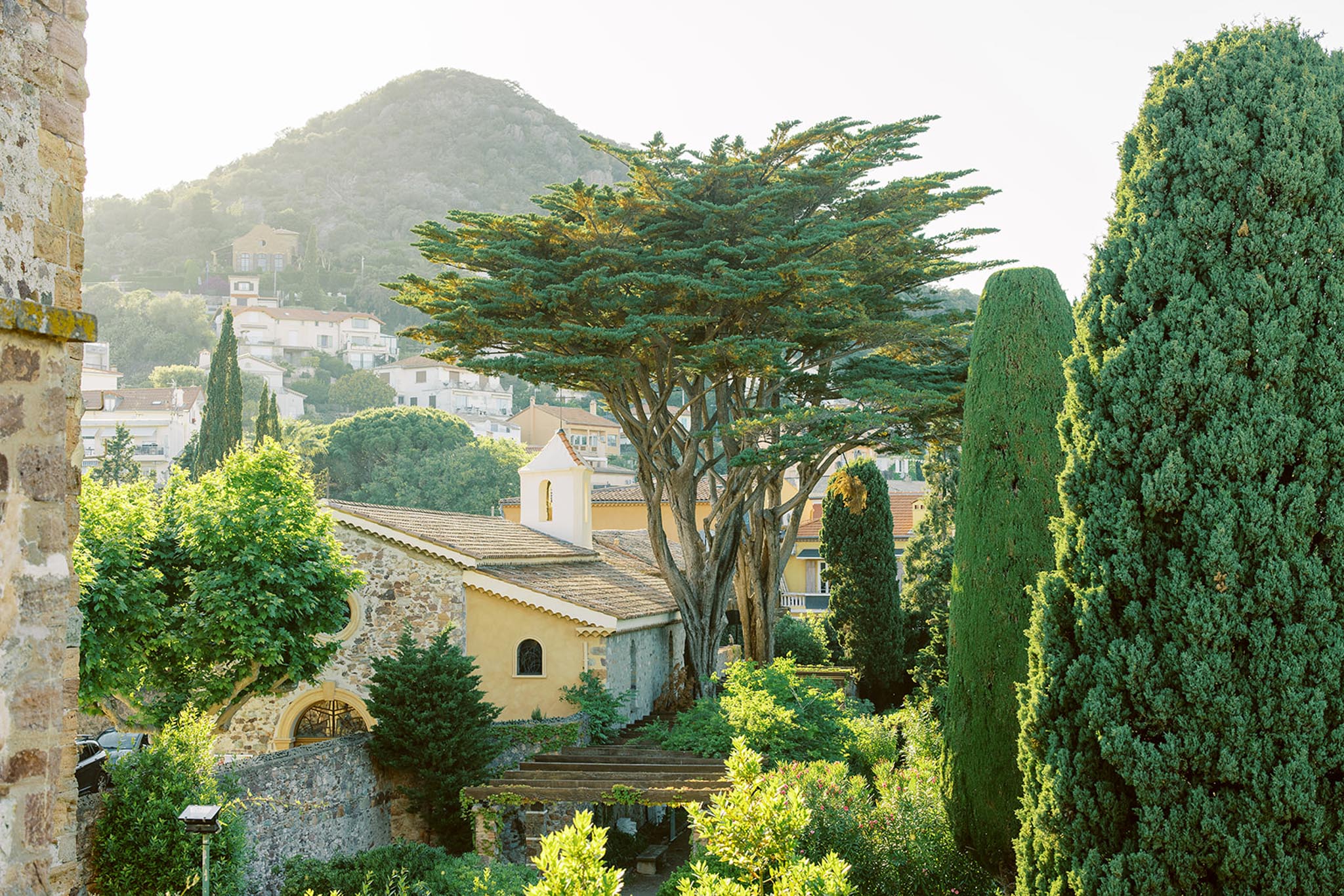 View over stone wall to yellow chapel with bell tower, cypress trees, and hillside village beyond
