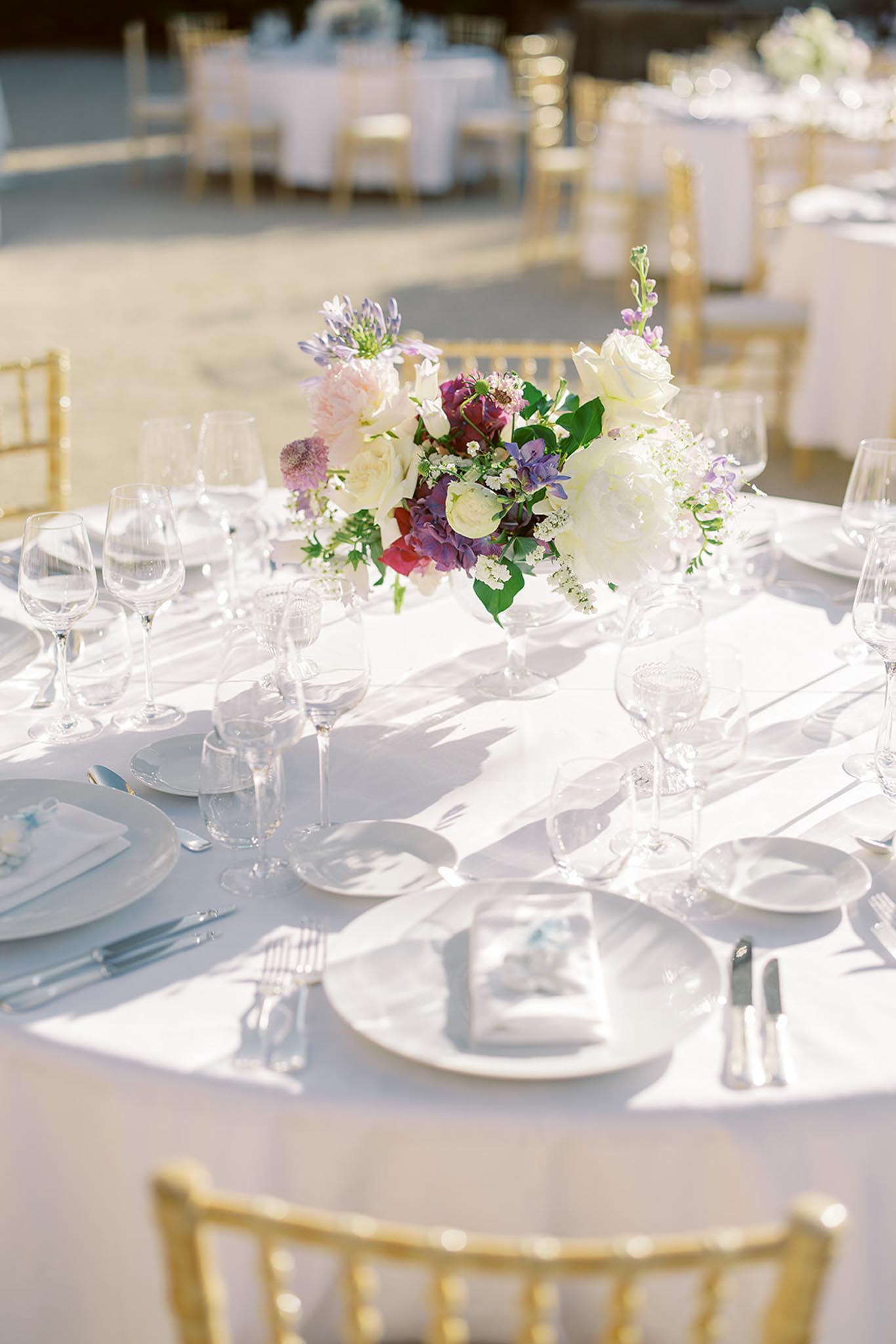 Round table with white and burgundy rose centerpiece, gold chiavari chairs, and crystal glassware in sunlight