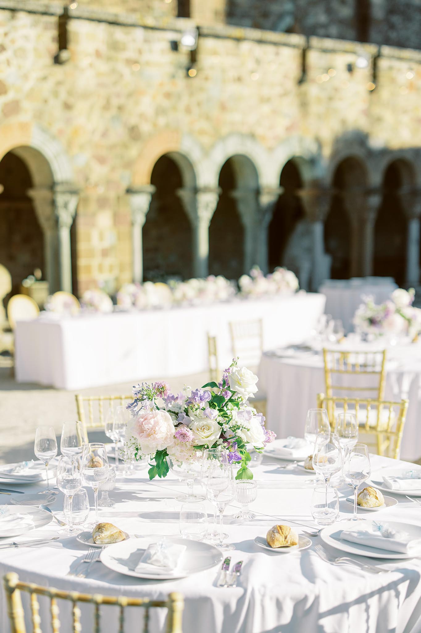 Round table with blush peony and lavender sweet pea centrepiece and gold Chiavari chairs in stone cloister