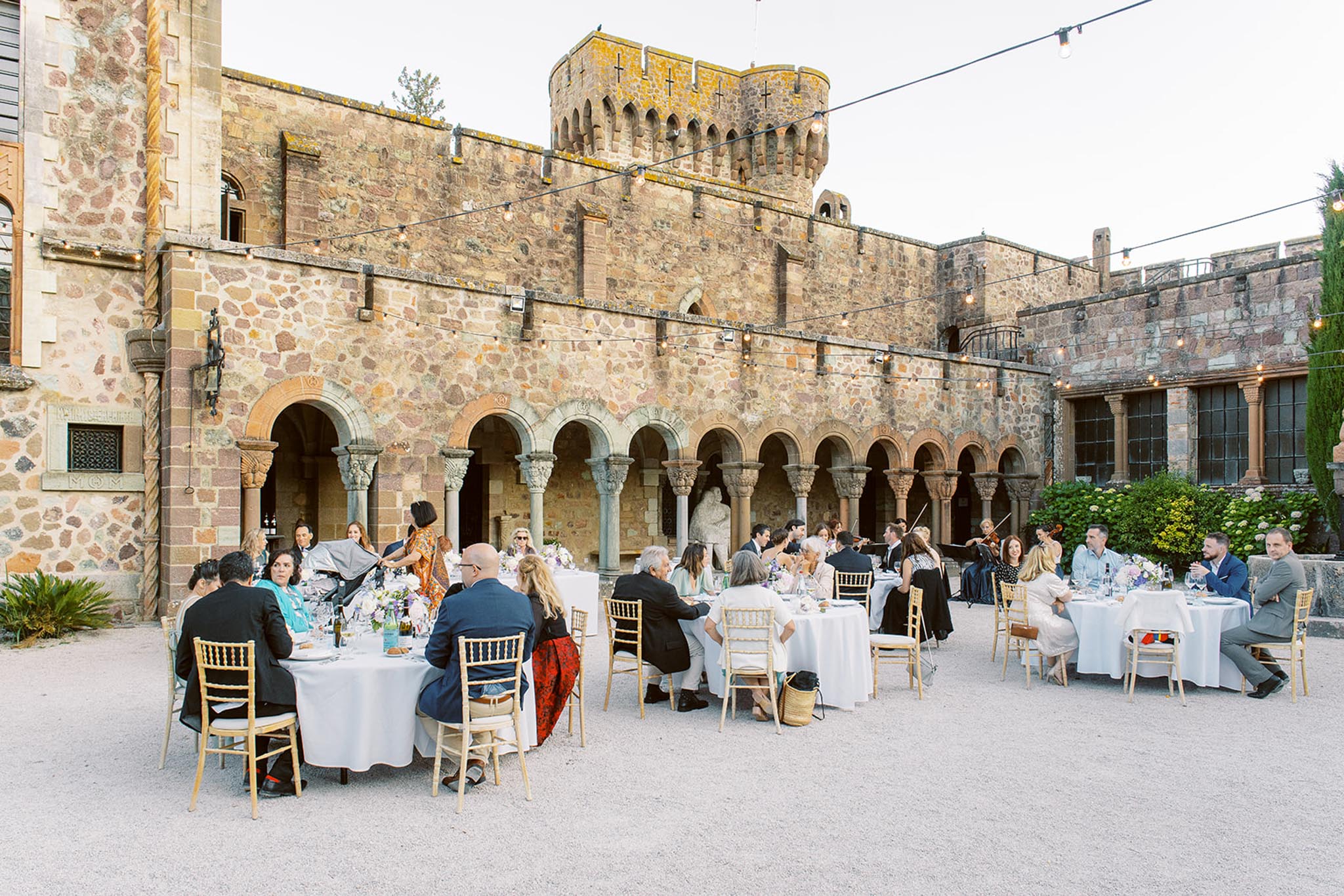 An outdoor wedding reception dinner is underway in the courtyard of a medieval-style castle or château, featuring a prominent round crenellated tower and an arched Romanesque cloister arcade along the facade. Approximately 30–40 guests are seated at round tables covered with white linen tablecloths, arranged across a gravel courtyard, with gold chiavari chairs throughout. Table centerpieces include loosely arranged floral arrangements in white, lavender, and soft purple tones. String lights are strung across the courtyard above the tables, providing warm ambient lighting in the early evening. Guests are dressed in smart-casual to formal attire in a mix of colors including navy, black, grey, and bright prints. What appears to be a string musician is visible performing near the cloister in the background. The overall styling is classic with relaxed details, set against the historic stone architecture. Wide establishing shot. Potential venue feature image.
