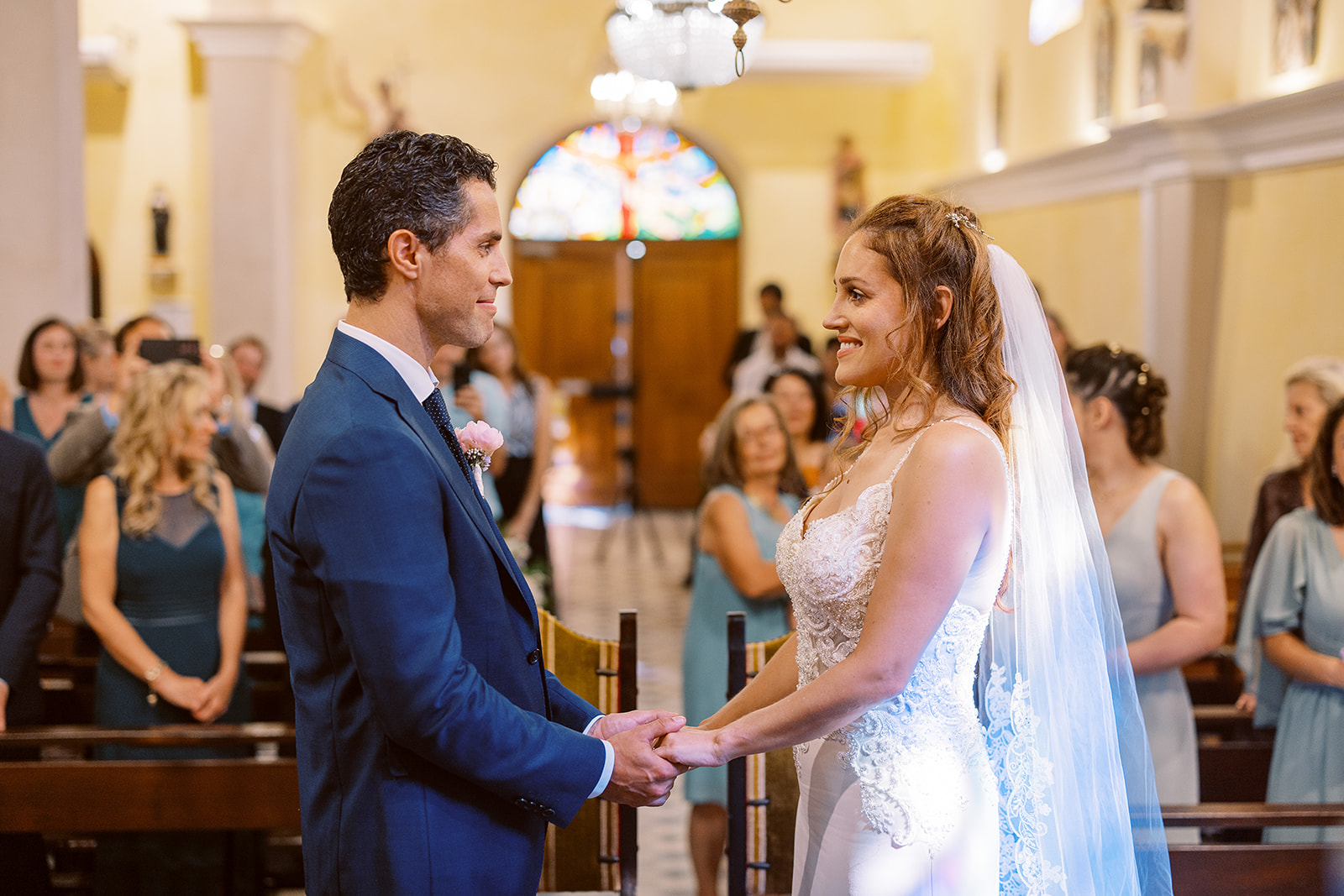 Bride and groom holding hands at church altar with stained glass window and guests standing in pews behind