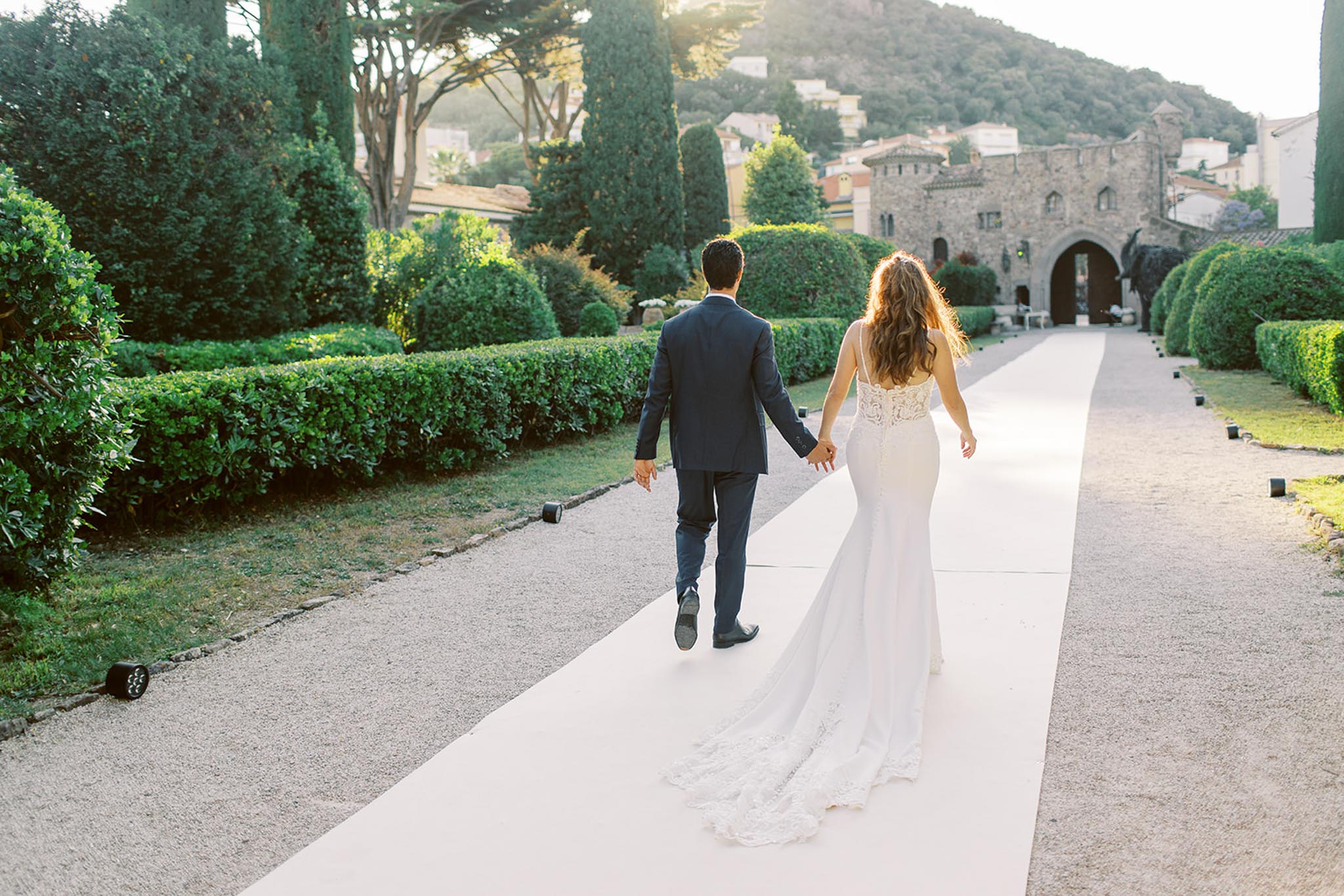 Bride and groom walk hand-in-hand toward medieval stone arches on cypress-lined path at golden hour