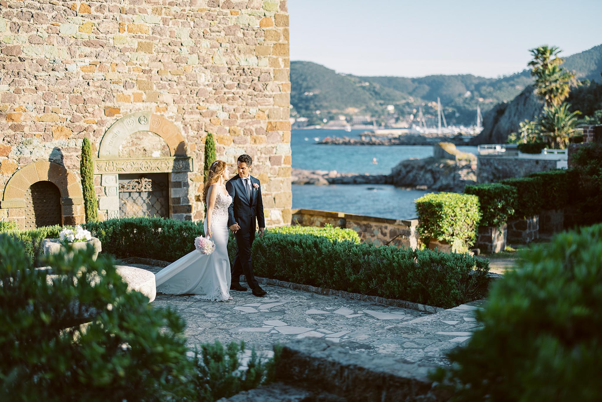 Couple walking on stone terrace with marina and hillside panorama at Mediterranean seaside venue