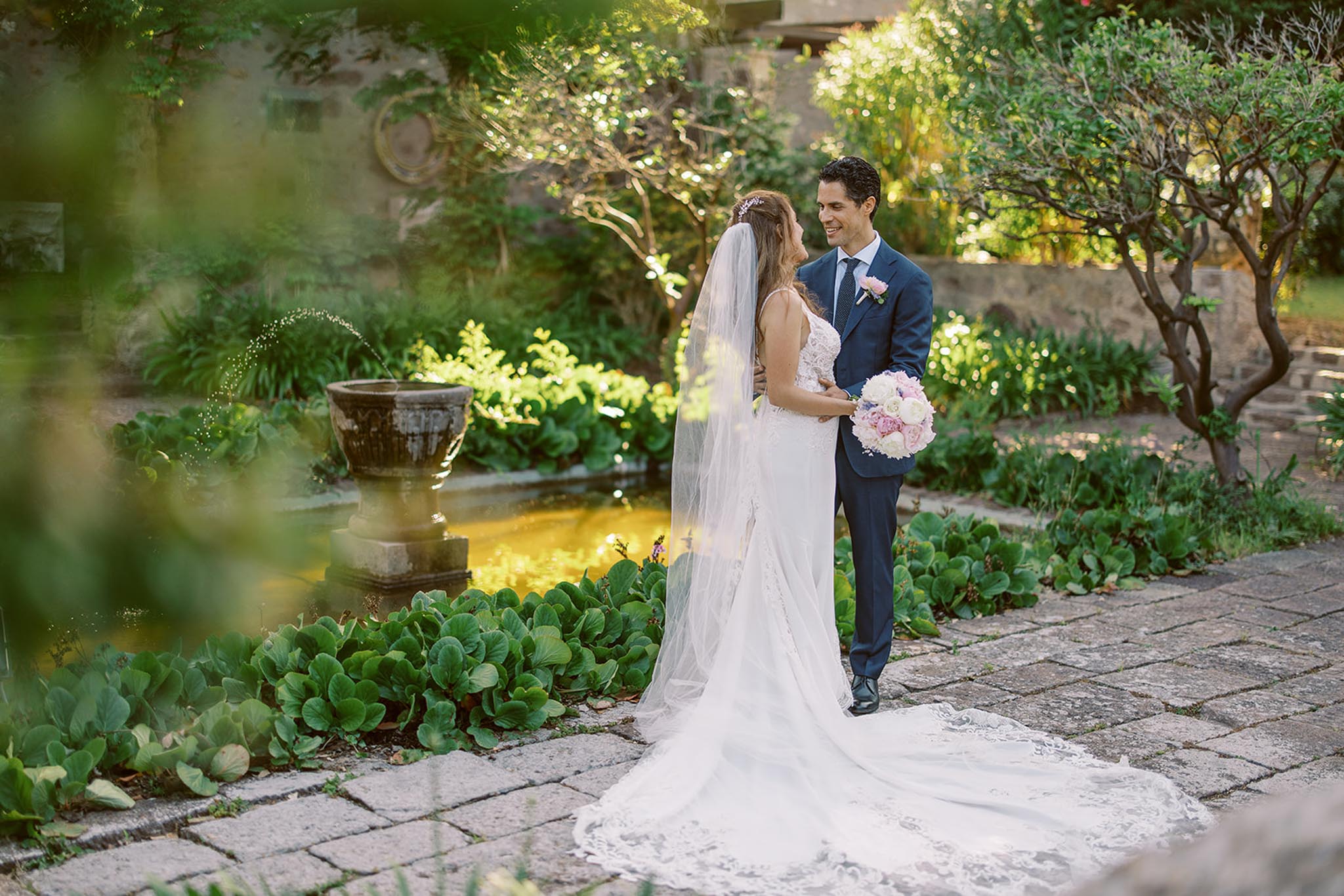 Bride and groom smiling in formal garden with stone fountain, bride holding blush peony bouquet
