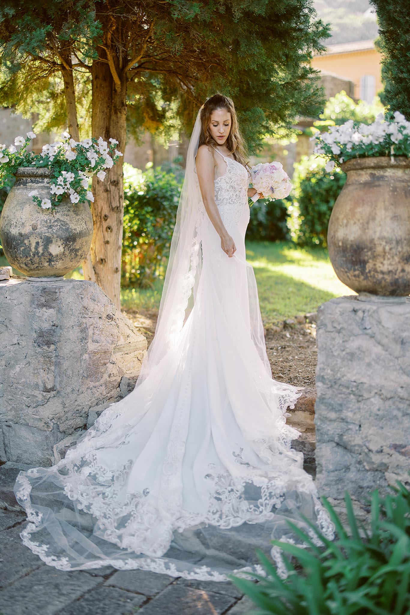 Bride in ivory lace gown with cathedral veil holding blush and lavender bouquet between terracotta urns in garden