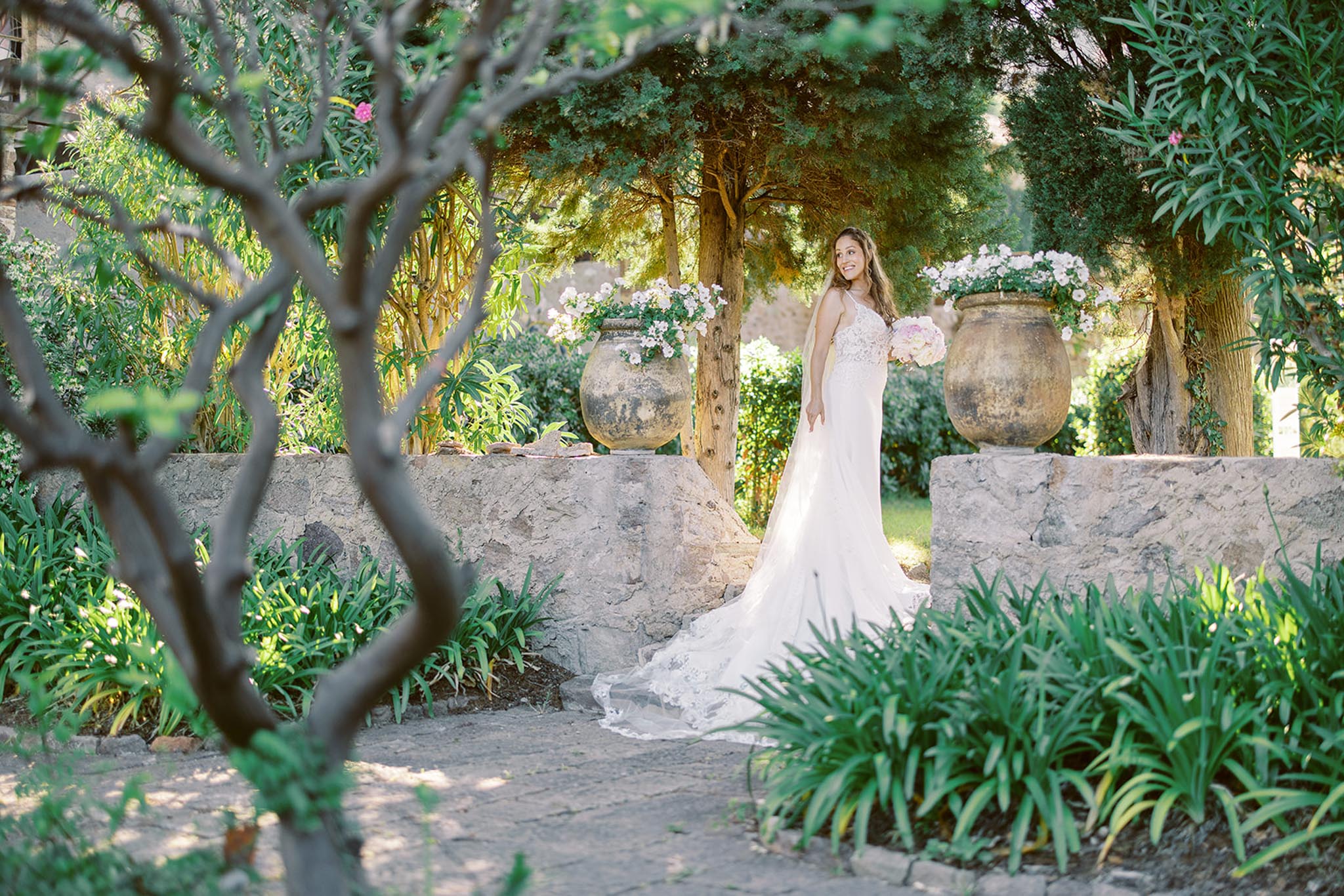 A bridal portrait taken outdoors in a formal garden setting, with the bride standing between two large weathered terracotta urns planted with soft pink and white flowers. The bride wears a fitted ivory lace gown with a spaghetti-strap bodice, a long cathedral-length lace-edged train, and holds a rounded bouquet of blush pink and white blooms. She is positioned beside a low rustic stone wall and looks slightly upward with a smile. The composition is a wide environmental portrait shot through foreground tree branches, which frame the scene and add depth. The overall styling aesthetic is classic Mediterranean, with the stone architecture and aged pottery contributing to a warm, sun-dappled garden atmosphere.