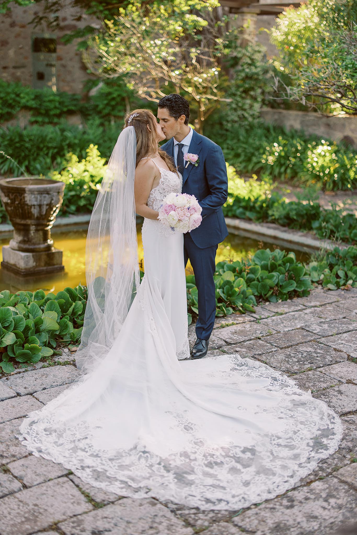 Couple kissing in walled garden bride in lace gown with cathedral veil and blush peony bouquet groom in navy suit