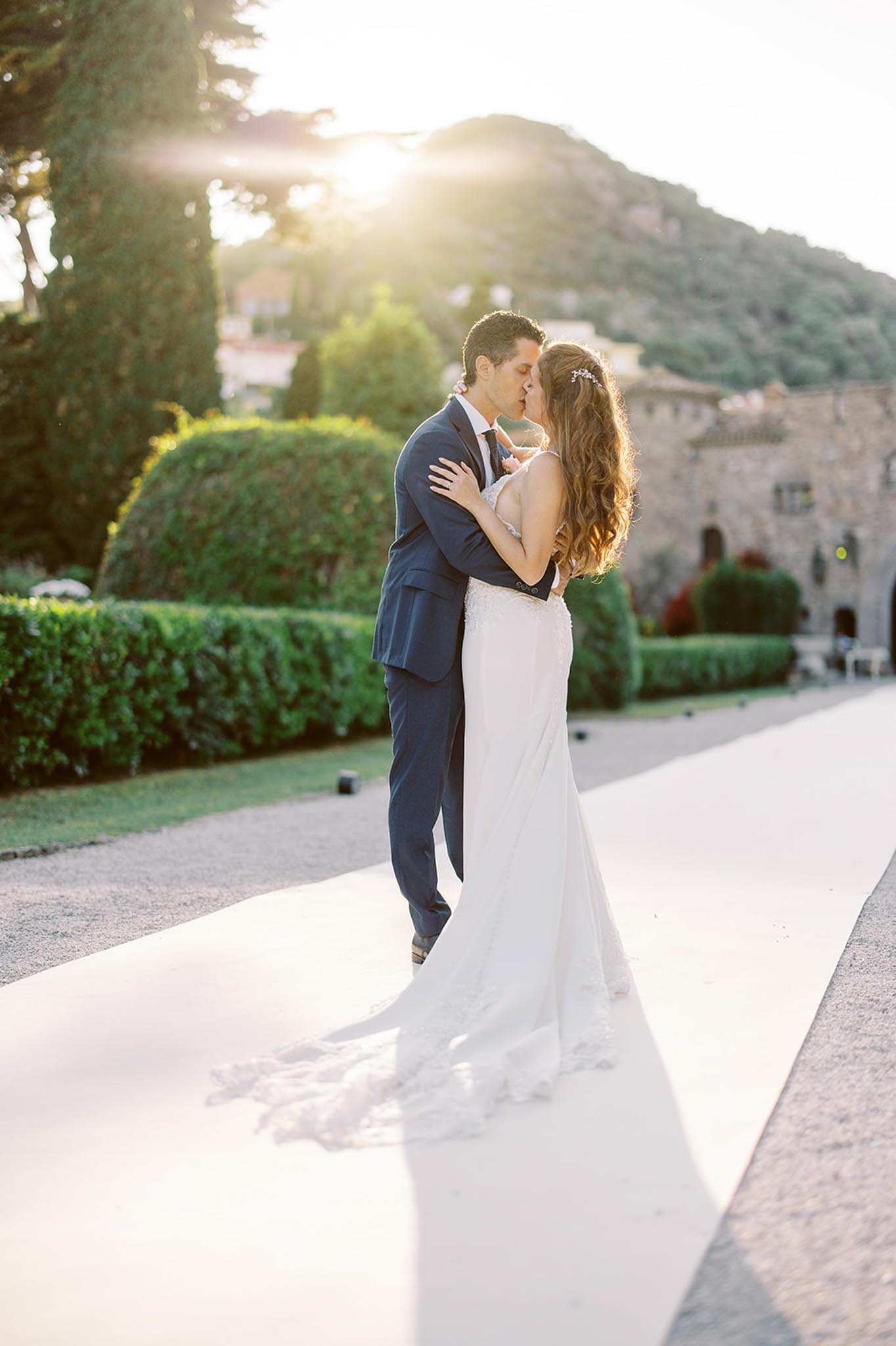 Couple kissing on paved path at golden hour with sun flare trimmed hedges and historic stone building behind