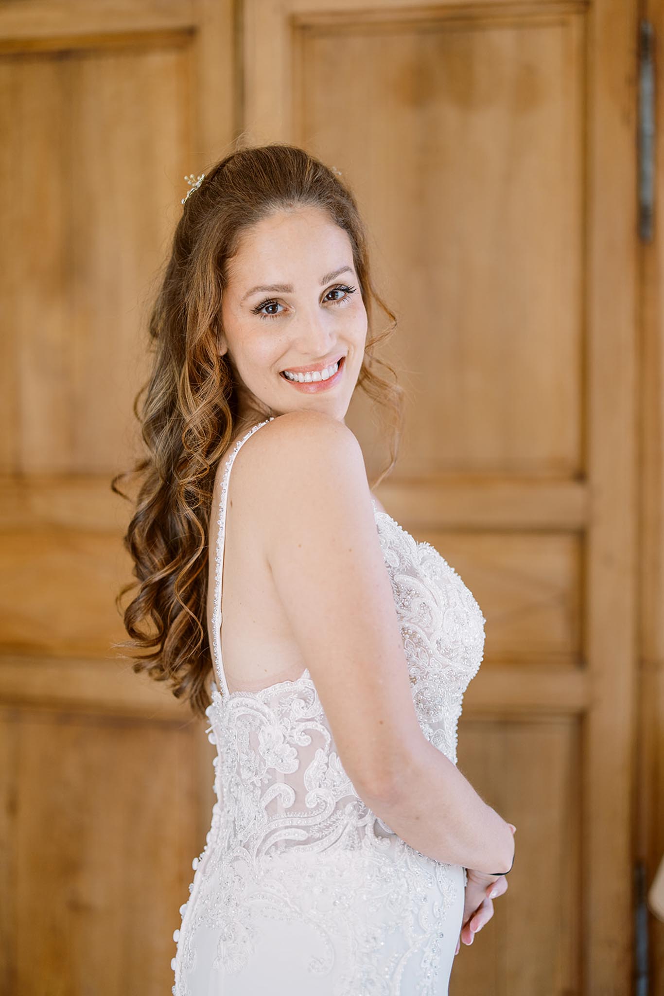 Bride looking over shoulder in fitted lace and beaded gown with open back before wooden panelled doors