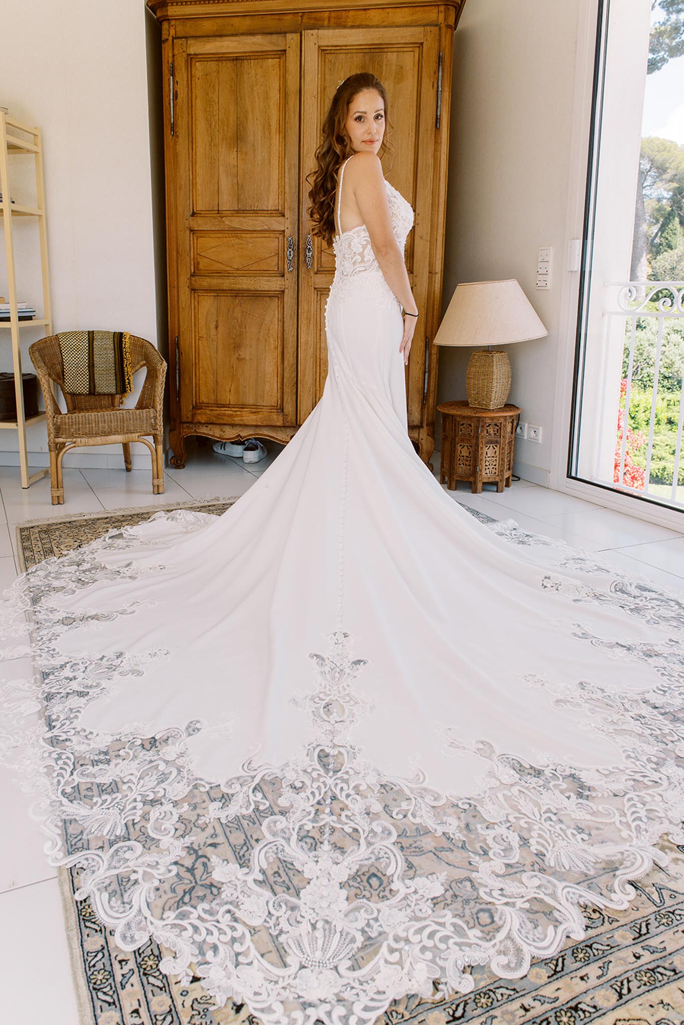 A bridal portrait taken indoors in a light-filled room, showing the bride standing and looking back over her shoulder toward the camera. She wears a fitted ivory mermaid-style wedding gown with thin spaghetti straps, lace appliqué bodice detailing, and an exceptionally long cathedral train edged with intricate scrolling lace embroidery. Her auburn hair is styled in loose waves. The room features white tiled floors, a large antique wooden armoire, a wicker chair with a patterned throw, a rattan side table with a lamp, and a floor-to-ceiling glass door opening to a balcony. The train fans out dramatically across a vintage-style blue and cream area rug, emphasizing its length and lace border detail. The composition is a full-length portrait shot from a slightly elevated angle to capture the full extent of the train.
