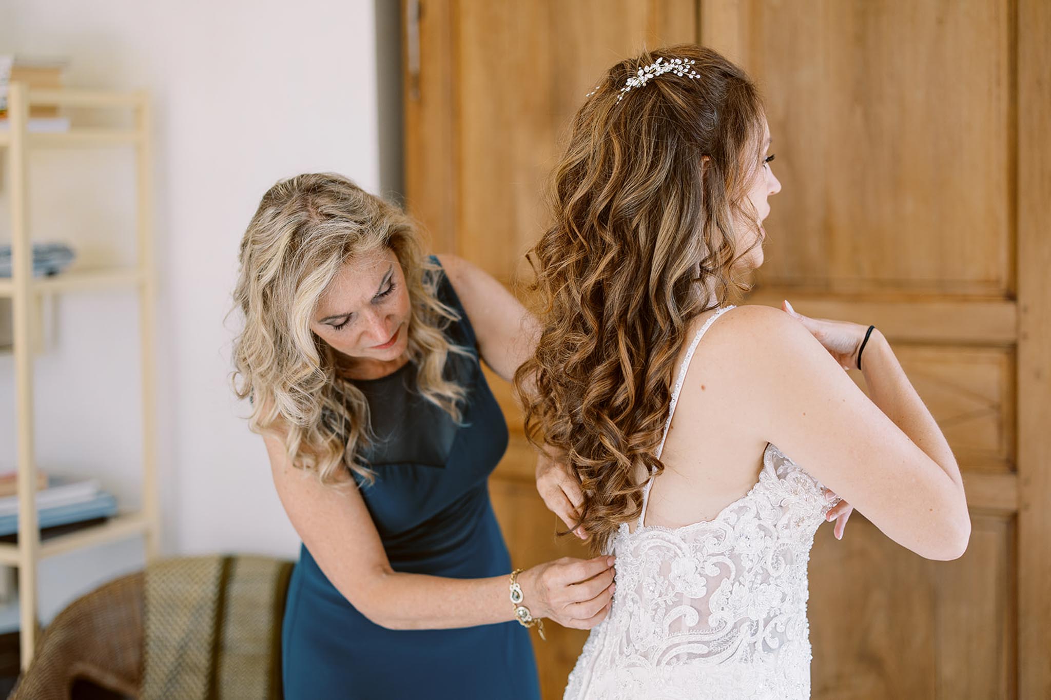 Mother fastening buttons on bride's low-back lace gown with crystal hair comb in warm-toned room