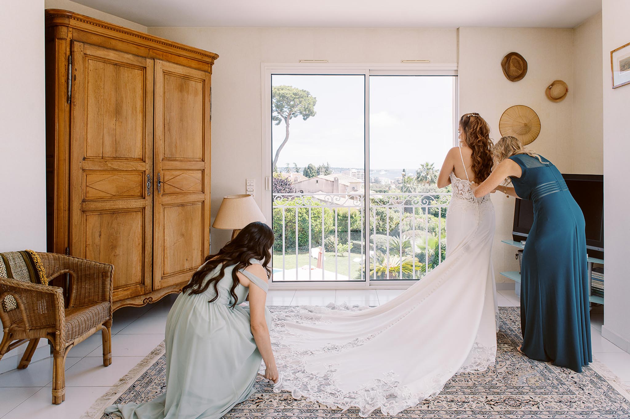 Two attendants in teal and sage dresses fastening bride ivory lace gown in Provencal bedroom