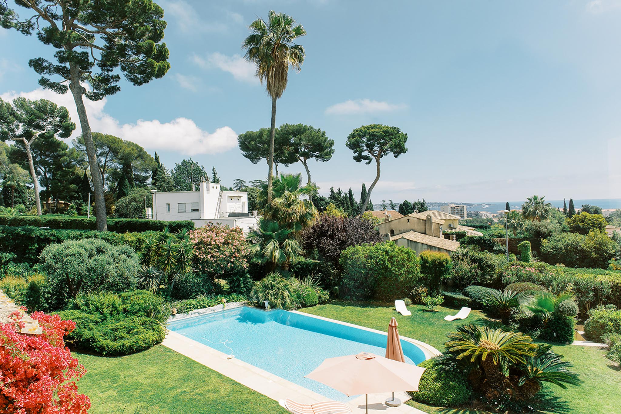 An elevated wide shot of a villa's outdoor grounds featuring a rectangular swimming pool with clear blue water, surrounded by a well-maintained garden. Two white sun loungers are positioned poolside to the right, and a closed peach-toned parasol with a matching base sits near the pool edge in the foreground. The garden is densely planted with a mix of palm trees, sculpted hedges, and flowering shrubs including red bougainvillea in the lower left. A stone-toned Provençal-style building is partially visible mid-frame, with a more modern white structure behind it, and a distant sea view is visible on the horizon to the right. No people are present in the image. Potential venue feature image.