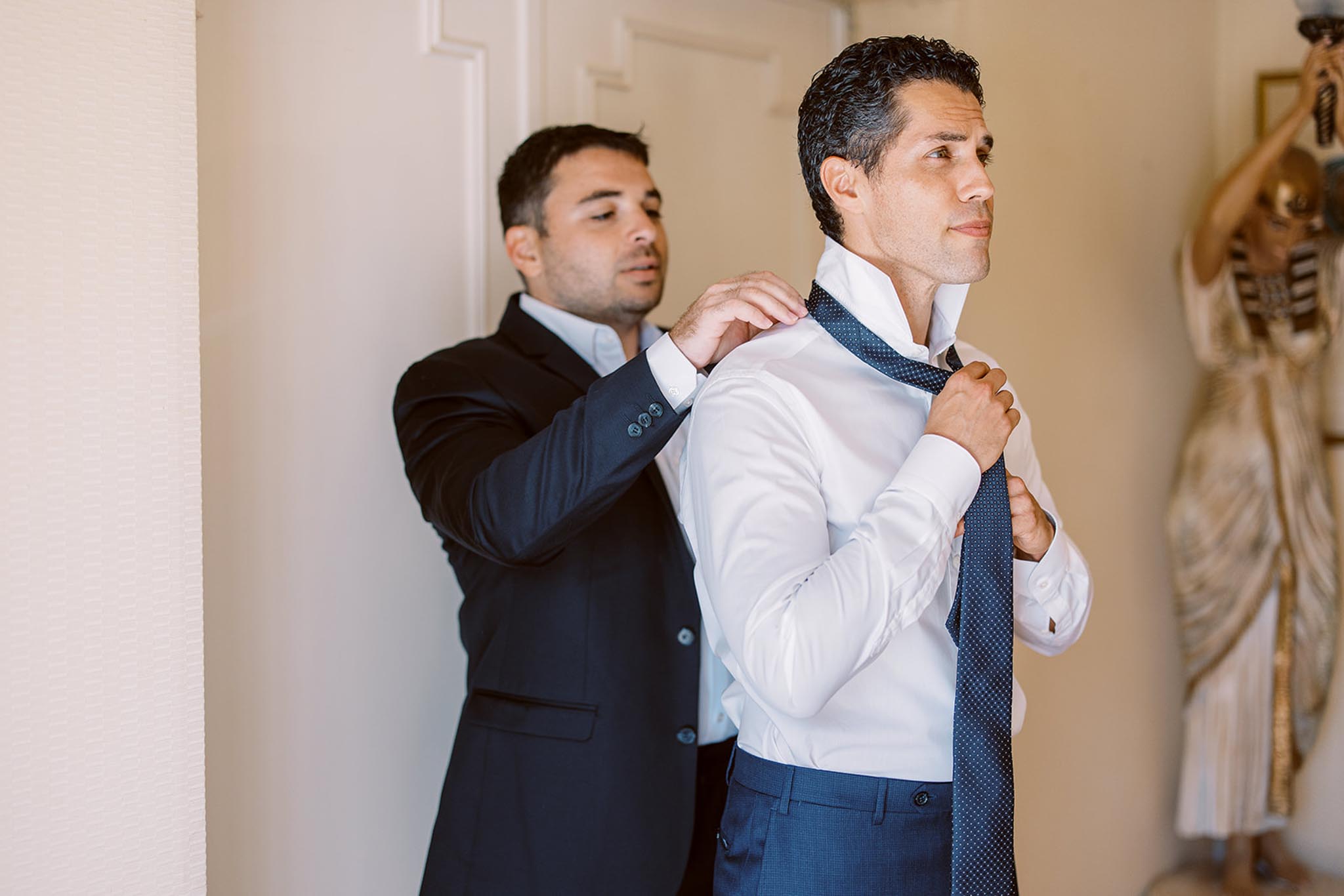 A getting-ready moment captured indoors showing two men, with one helping the other adjust a navy blue polka-dot tie. The groom is wearing a white dress shirt and navy blue trousers, while the man assisting him wears a dark navy suit jacket over a light blue shirt. The setting appears to be inside a venue with cream-colored walls, and a decorative gold and cream figurine sculpture is partially visible in the background to the right. The shot is a medium portrait framing both men from approximately the waist up, with natural light illuminating the scene from the left.