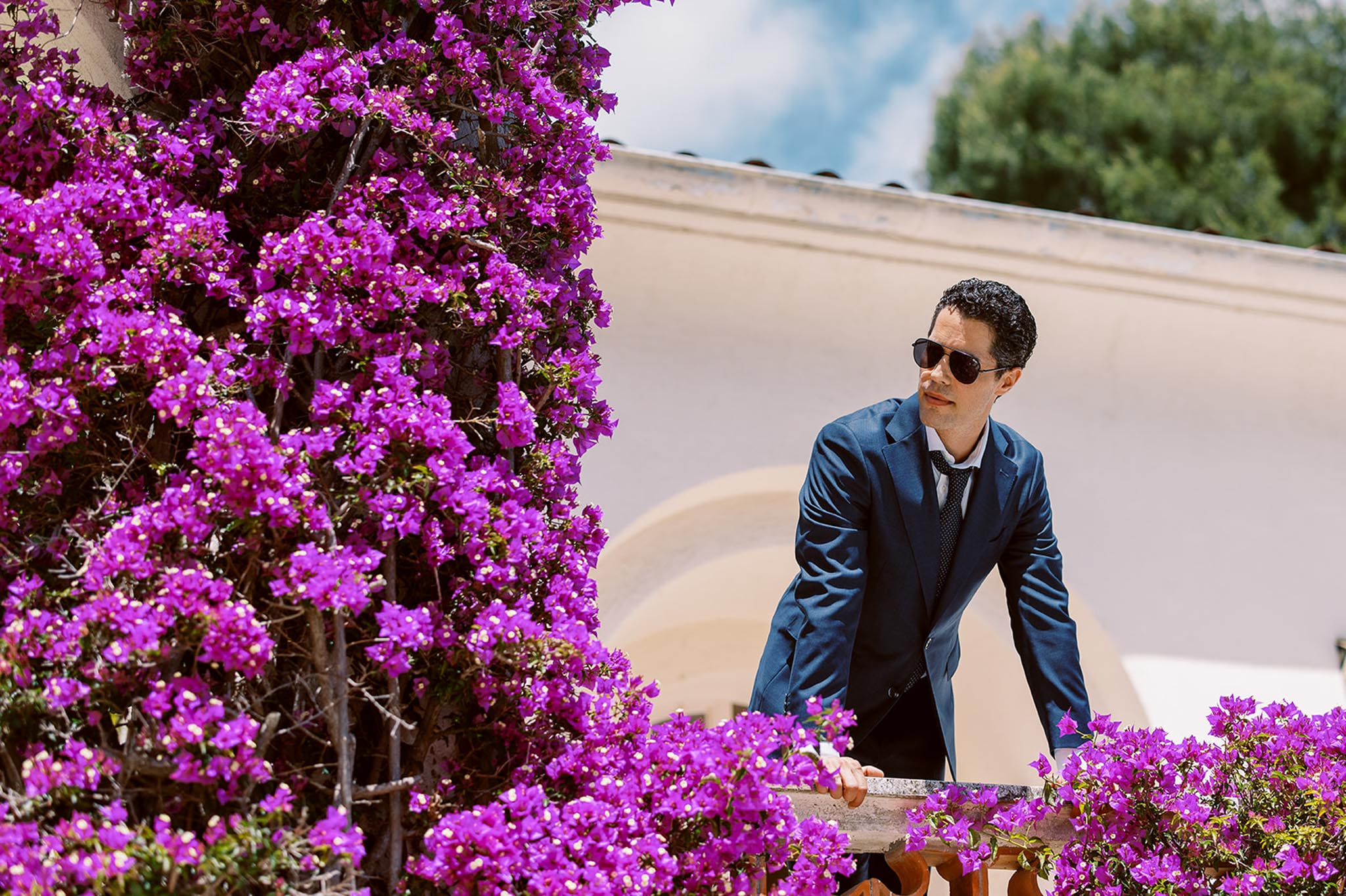 Groom in navy suit and sunglasses leaning against stone balustrade with magenta bougainvillea at Mediterranean venue