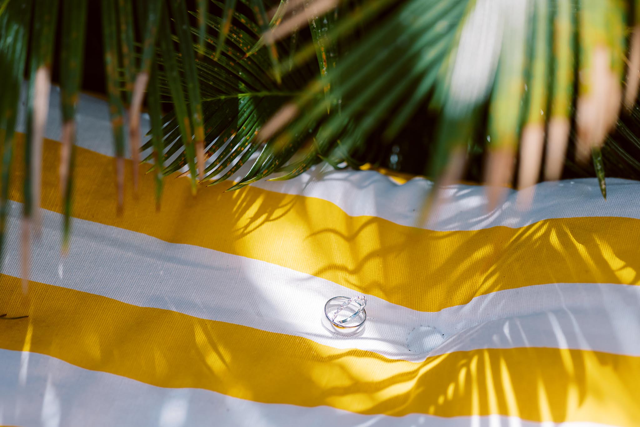A close-up detail shot of two wedding rings — one plain silver band and one silver ring with small diamonds — placed on a yellow and white striped fabric surface, likely a sun lounger or cushion. Palm fronds hang over the top of the frame, casting dappled shadows across the striped textile. The composition frames the rings at the lower center of the image, with strong natural sunlight creating high contrast shadows from the palm leaves. The styling suggests a relaxed, tropical or summer outdoor setting.