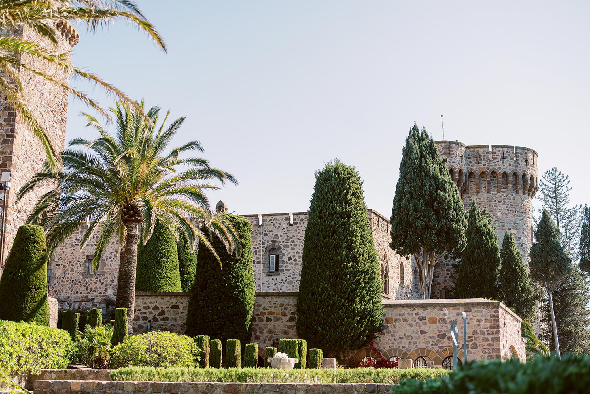 Medieval chateau with round crenellated tower, manicured topiary hedges, cypress trees, and palm tree on grounds