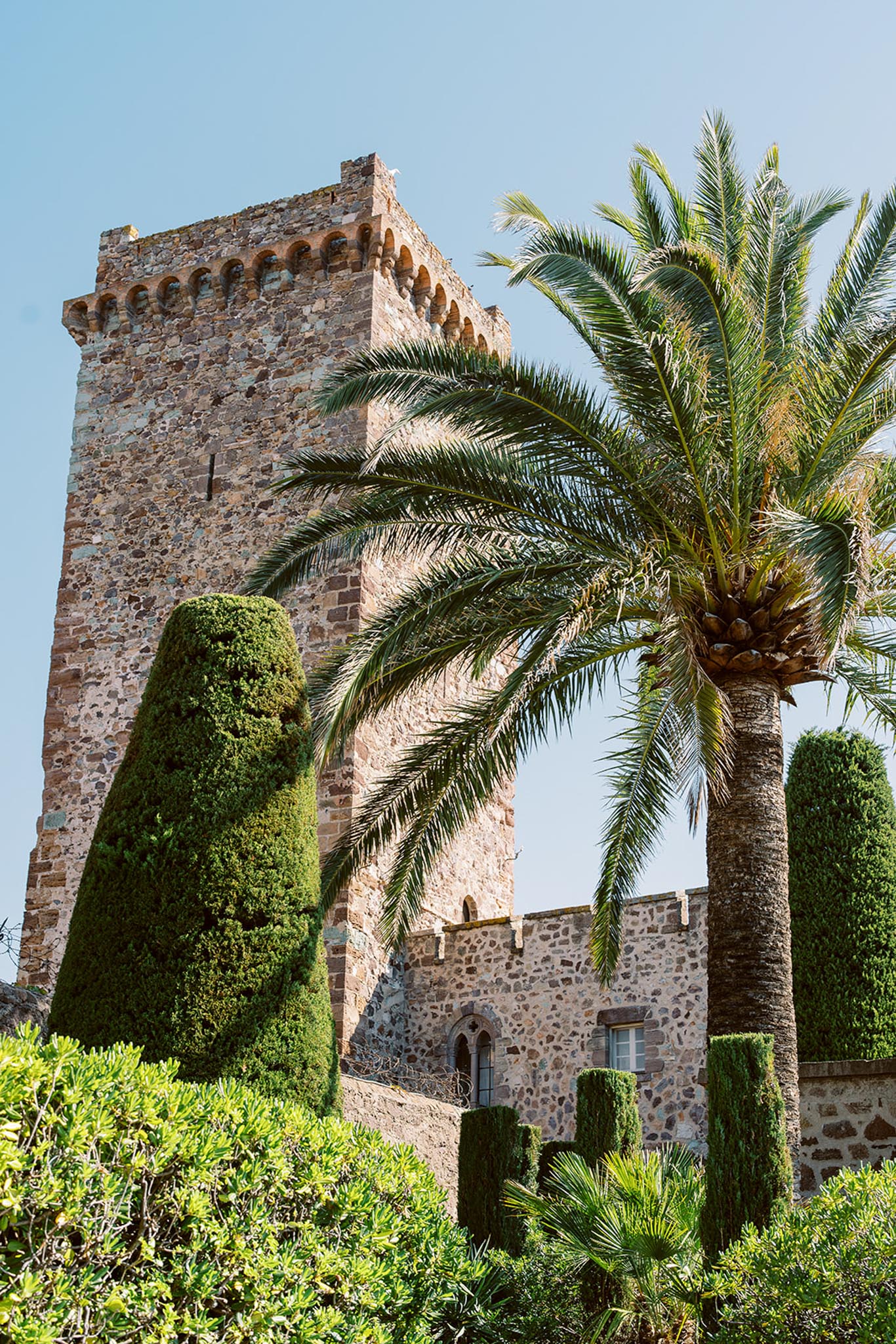 Medieval stone tower with crenellated battlements Romanesque arches conical cypress topiaries and mature palm tree