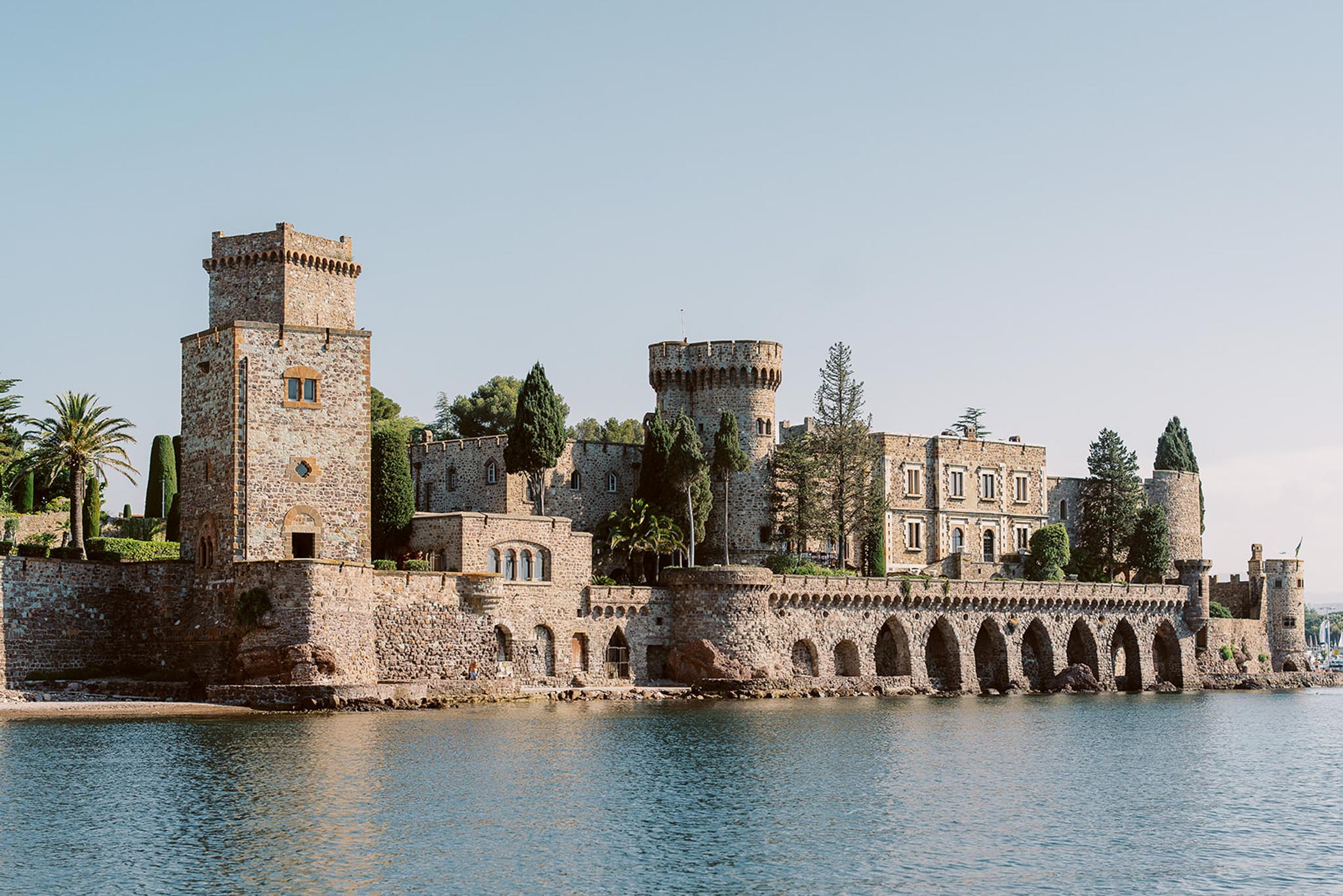Medieval waterfront castle with square tower, round turret, and arcaded stone wall reflected in calm water