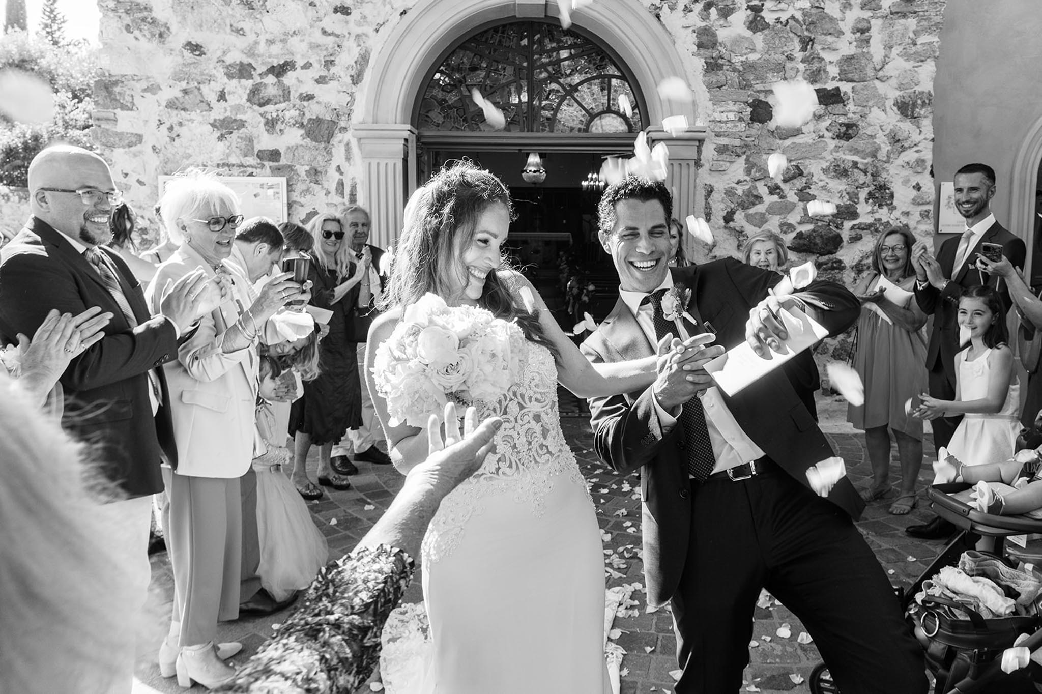 Black and white candid of couple exiting stone chapel as guests throw flower petals