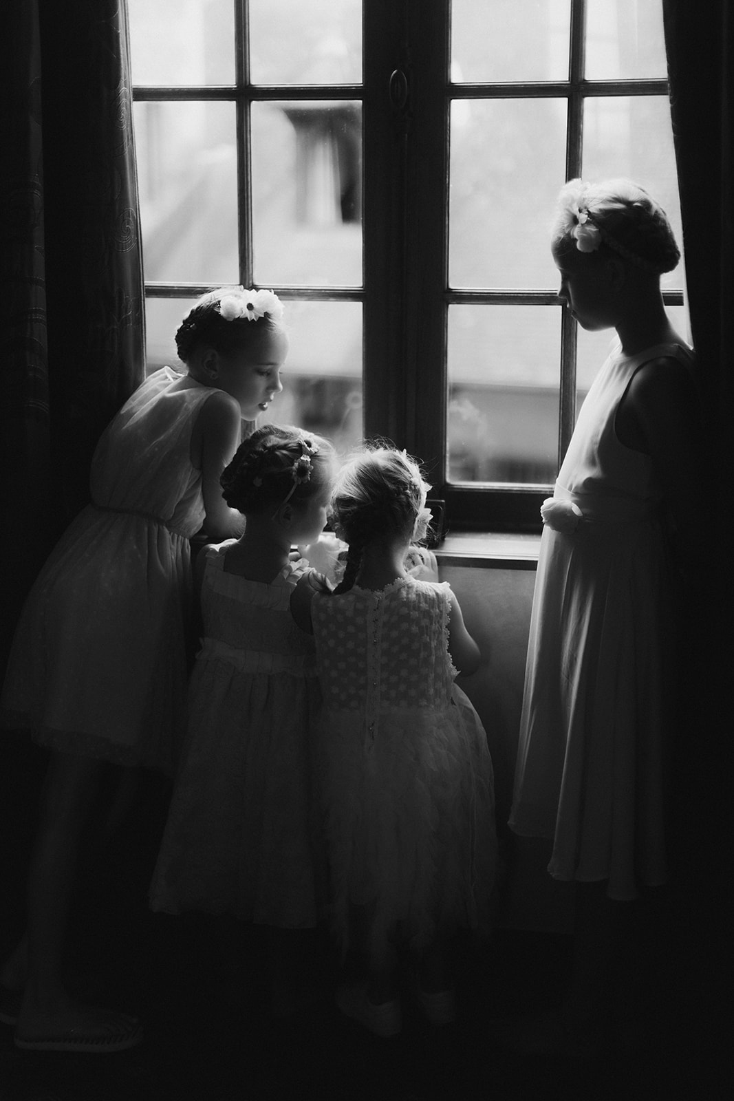Black-and-white image of four flower girls in white dresses gathered at a French window inside a chateau