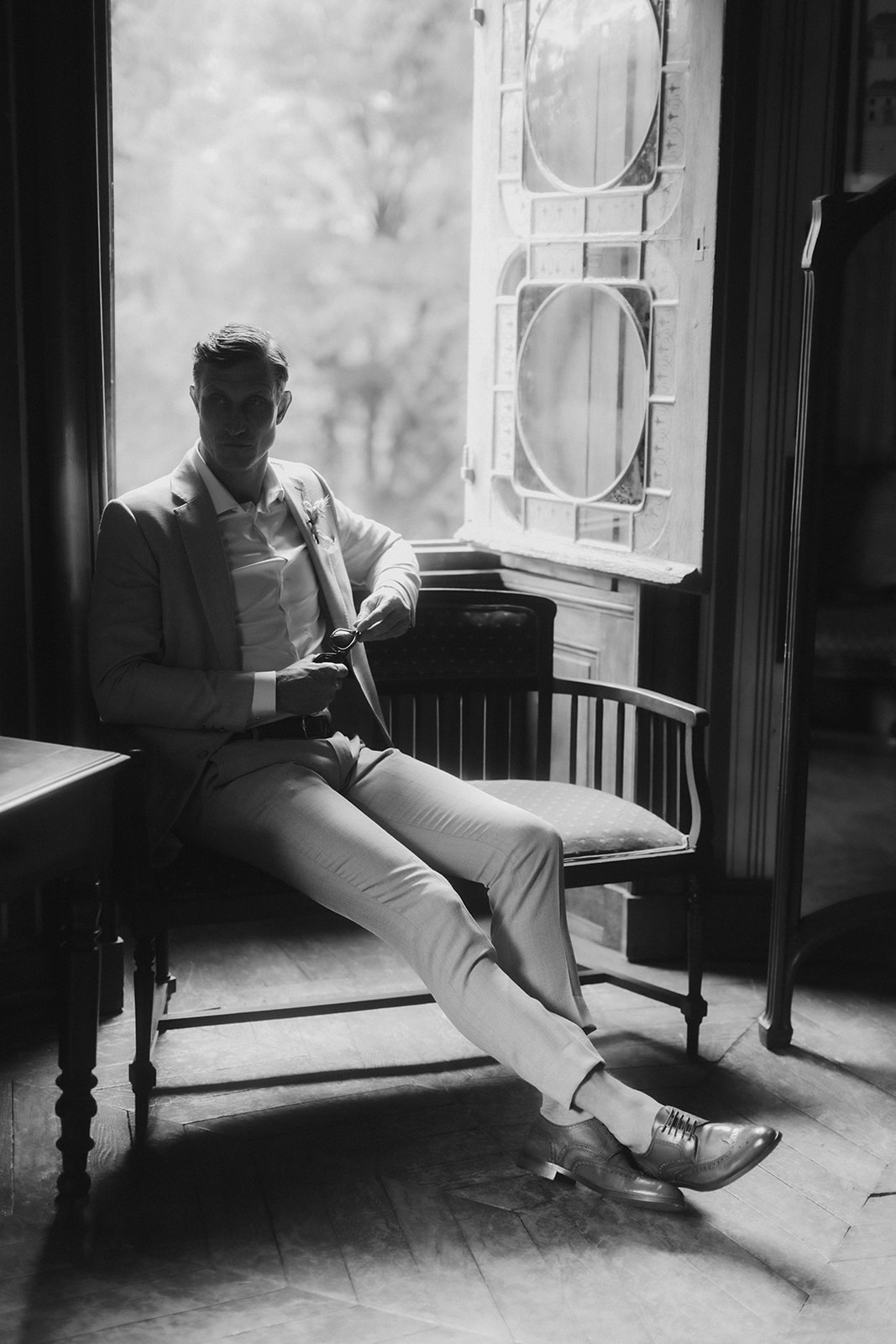 Black and white groom in light suit seated by leaded-glass window with rim-light effect in chateau parquet room