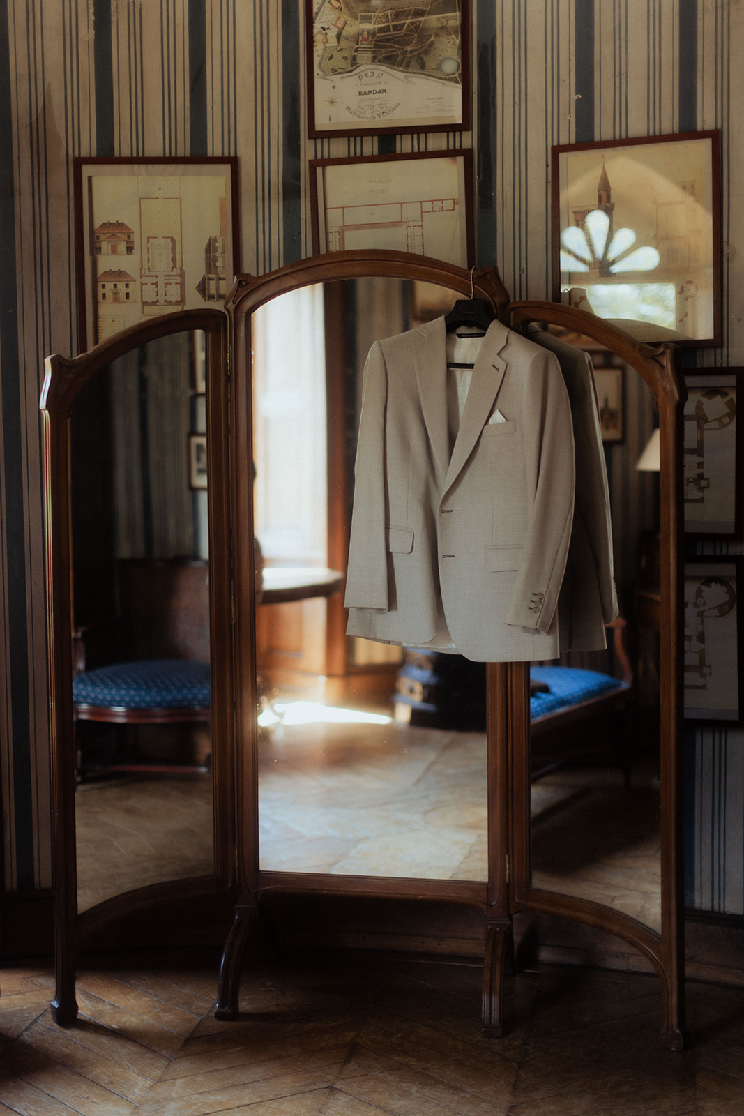 A groom's getting-ready detail shot taken indoors in a classically decorated room. A light grey suit jacket with a white pocket square is hung on a hanger over the top of a dark walnut antique three-panel dressing mirror. The room features blue and white striped wallpaper, parquet flooring, antique architectural prints and maps in wooden frames on the walls, and blue upholstered chairs visible in the mirror's reflection. Warm natural light streams through a window reflected in the central mirror panel. The overall interior styling is classic French period decor. Close-up to medium detail shot with the trifold mirror as the compositional anchor.