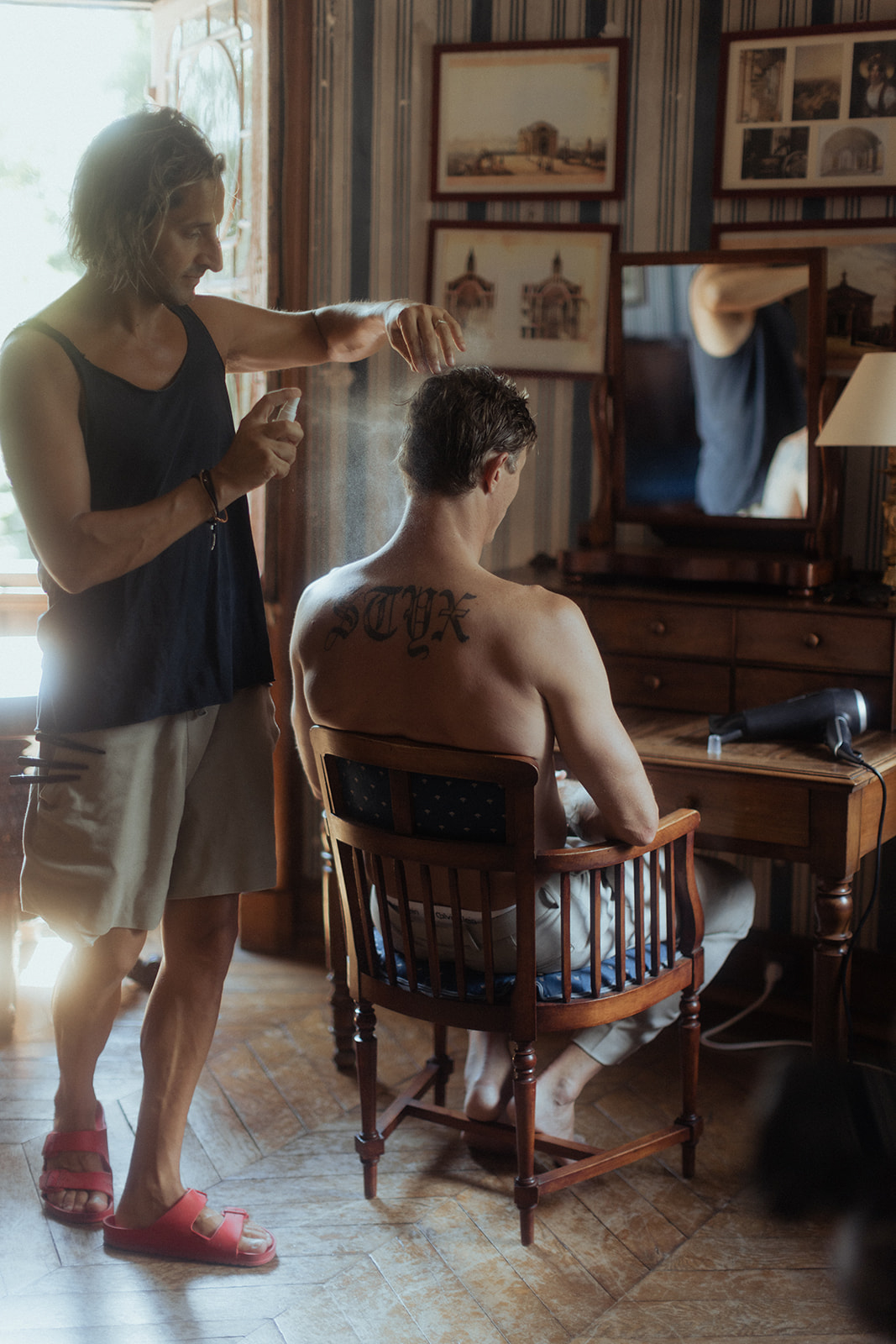 A getting-ready scene shot indoors, showing a shirtless man seated in a wooden chair while a stylist applies hair product via a spray bottle. The seated man has a large old-English lettering tattoo reading 'STYX' across his upper back. The room has a classic, lived-in feel with striped wallpaper, framed architectural prints on the walls, a wooden dressing table with a mirror and hairdryer visible in the background, and herringbone parquet flooring. Warm natural light streams through a window to the left, creating a hazy, backlit quality. The image is a medium full-length portrait shot.